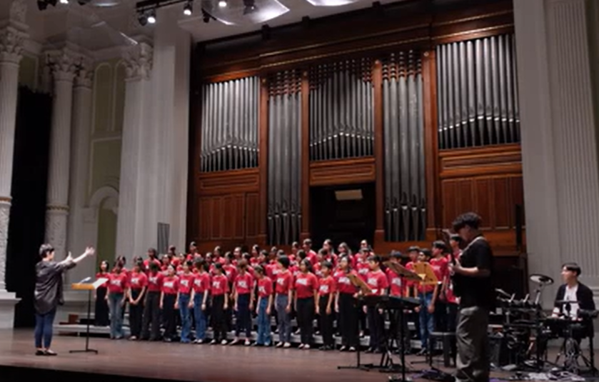 A children's choir in red shirts performs on stage before a large pipe organ, led by a conductor.