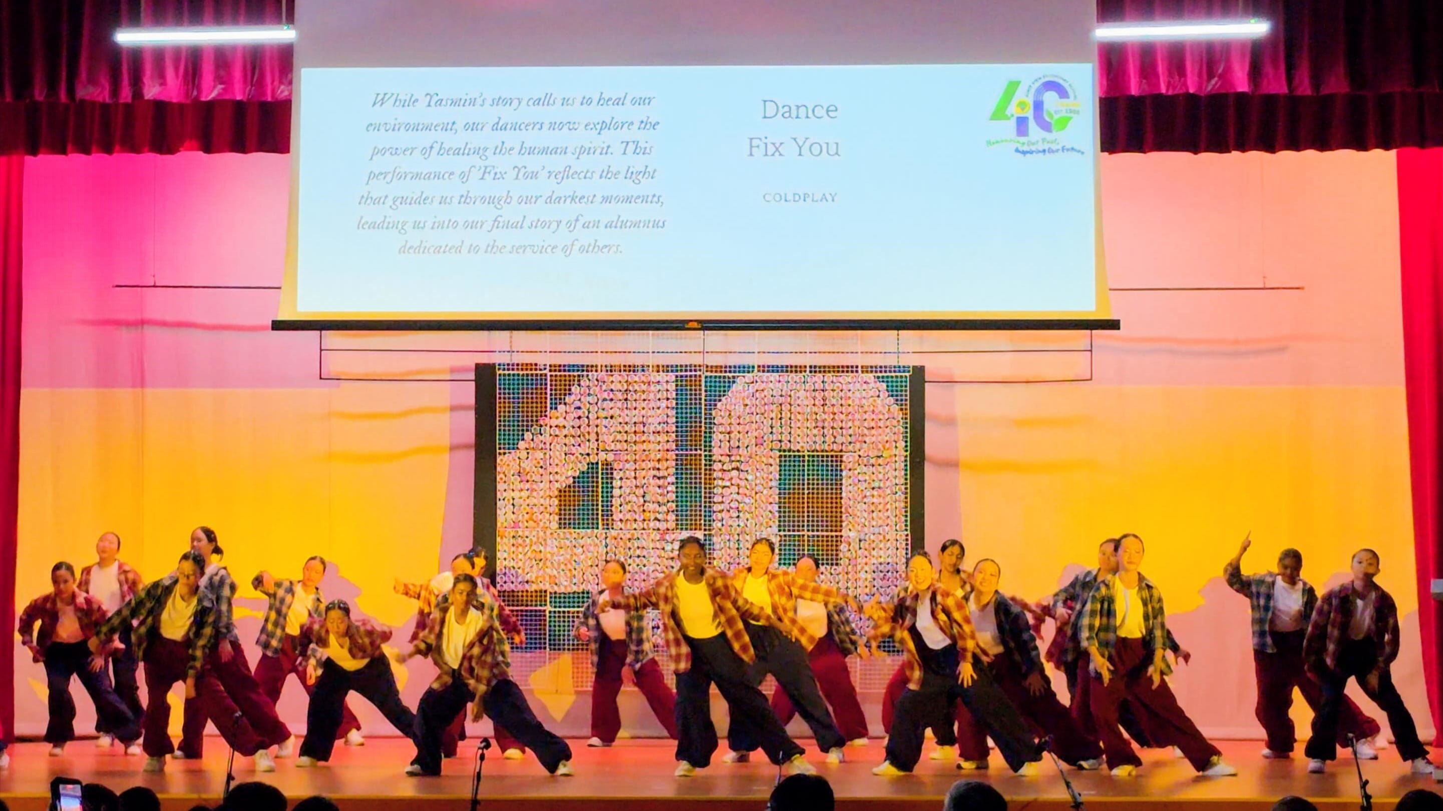 A large group of dancers in plaid shirts perform on stage to Coldplay's Fix You at a 40th anniversary event.
