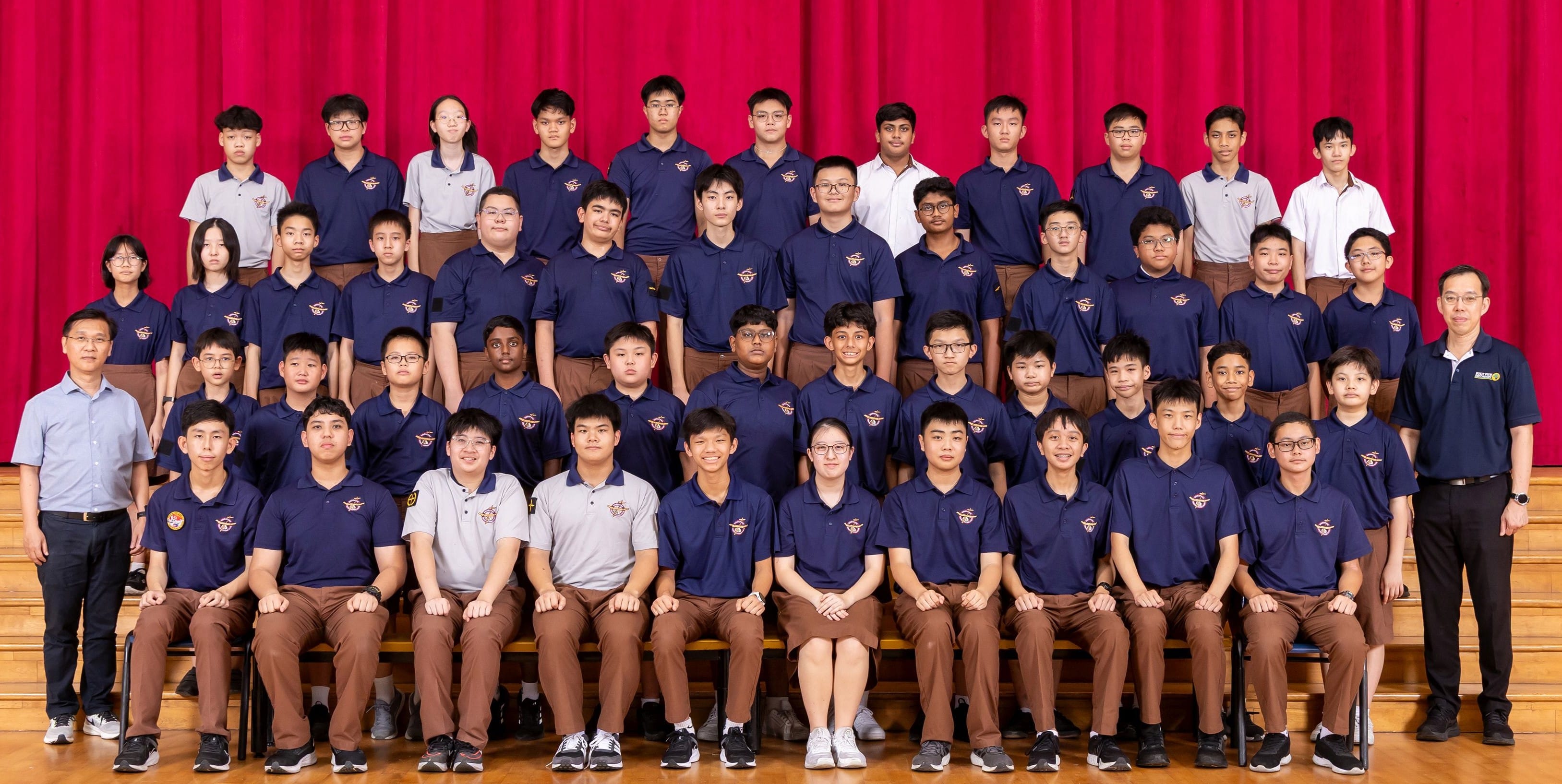 Large group of students in navy polo uniforms posed in rows on a stage with a red curtain backdrop.