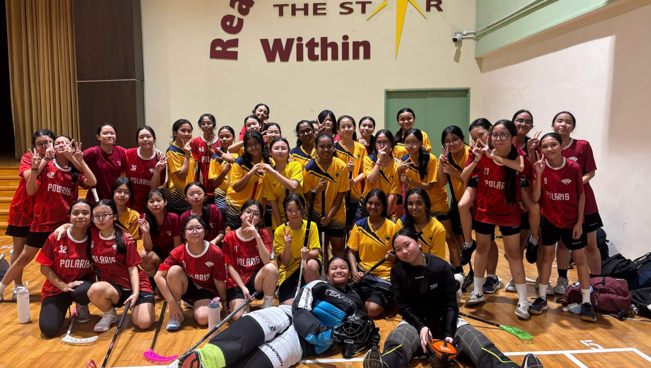 Group of girls in red and yellow sports jerseys posing in a gym, with floorball sticks and goalie gear.