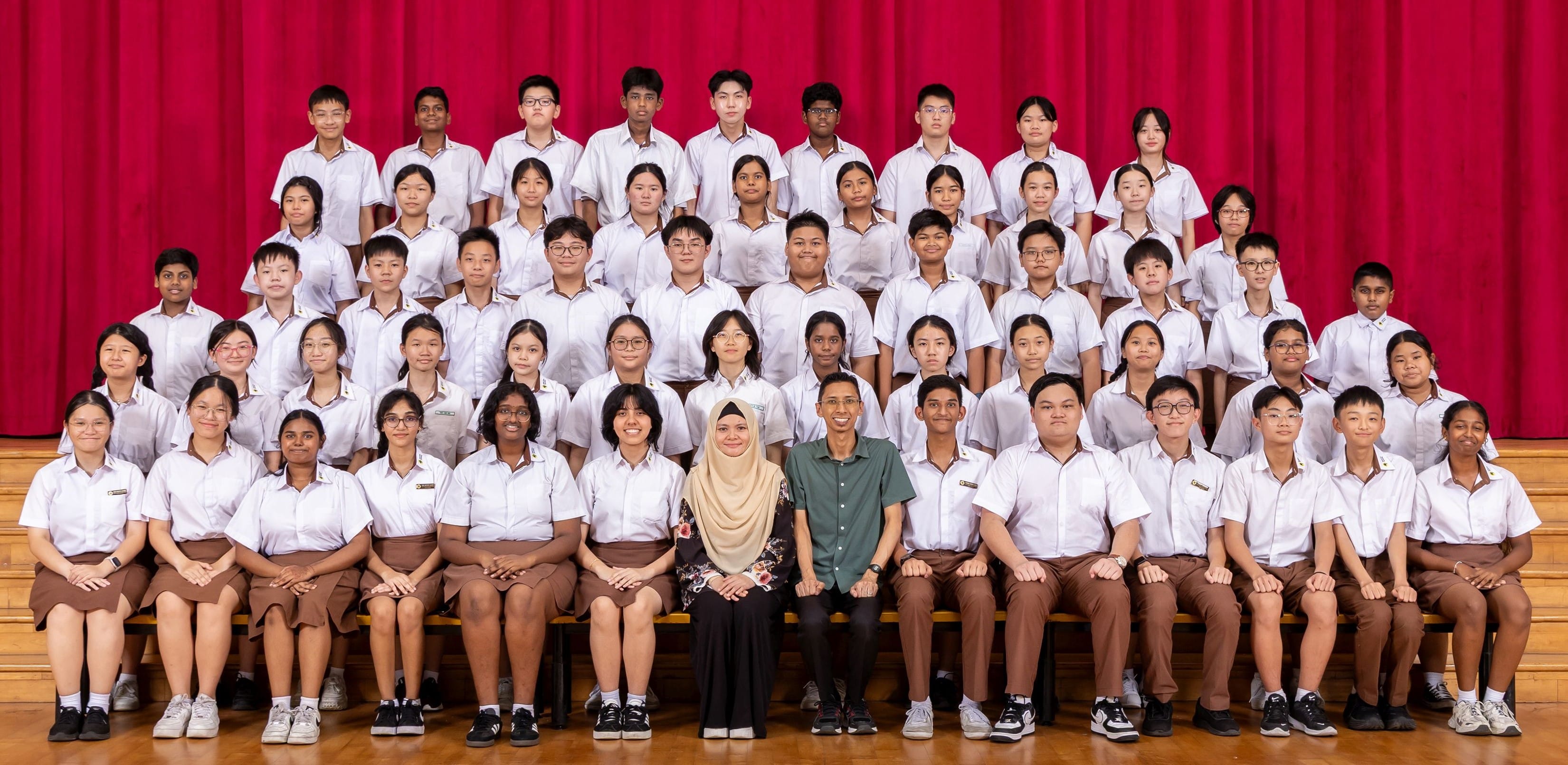 Large school group photo with students in white uniforms and two teachers seated in front of a red curtain.