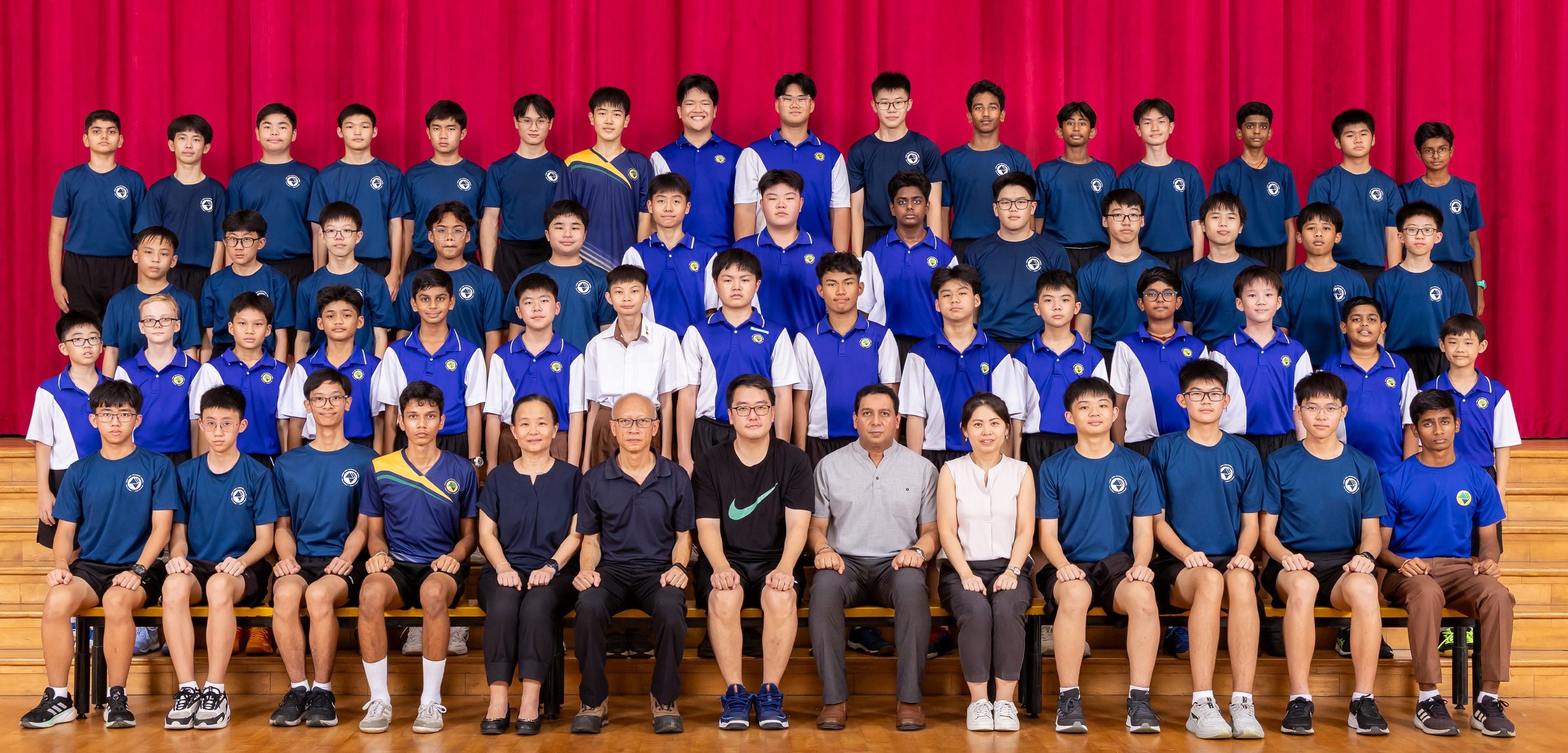 Large group of school boys in blue uniforms posed in rows with several adult teachers in front of a red curtain.