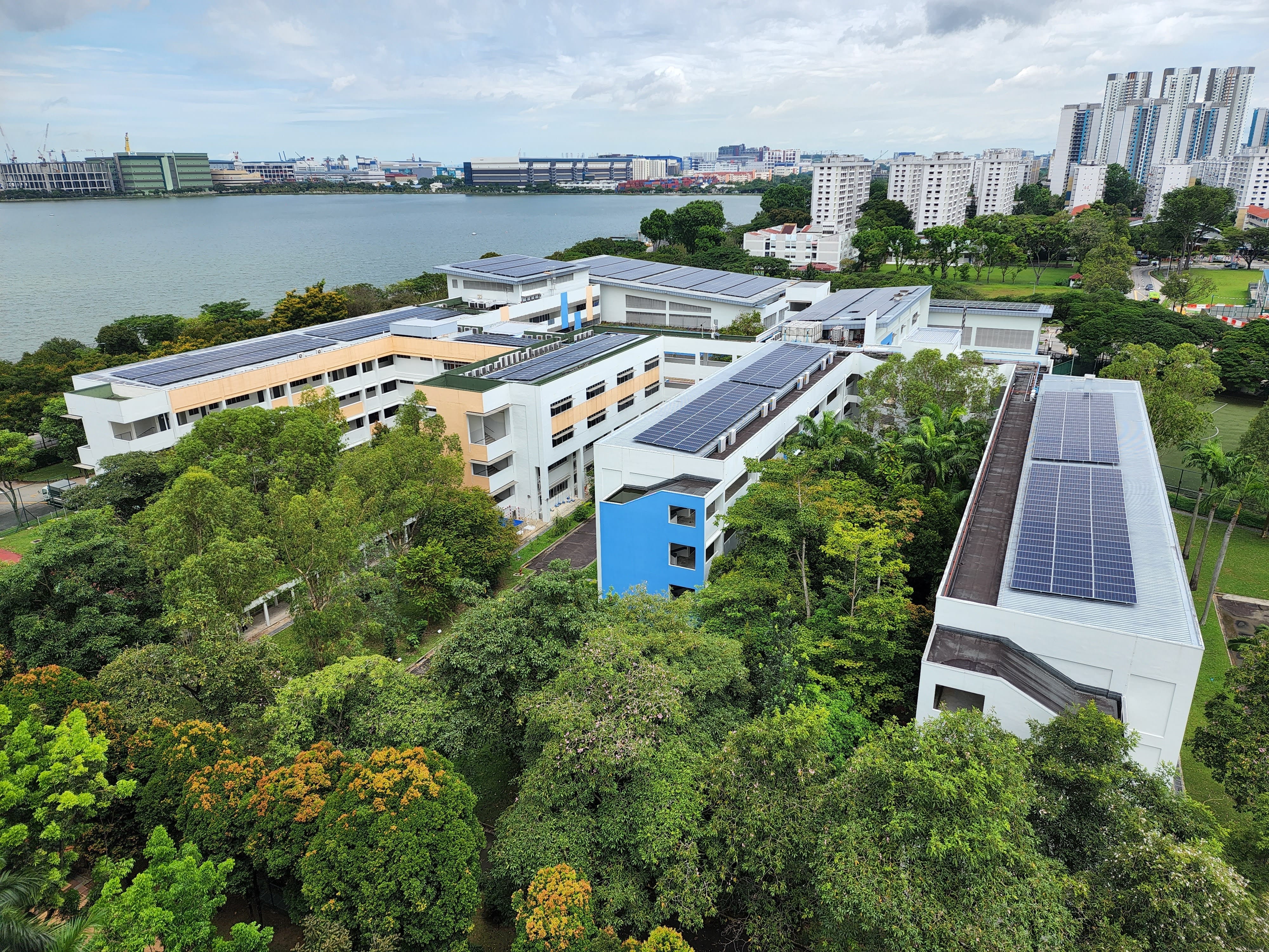 Aerial view of white multi-story buildings with solar panels on rooftops, surrounded by trees, near a waterway and city skyline.