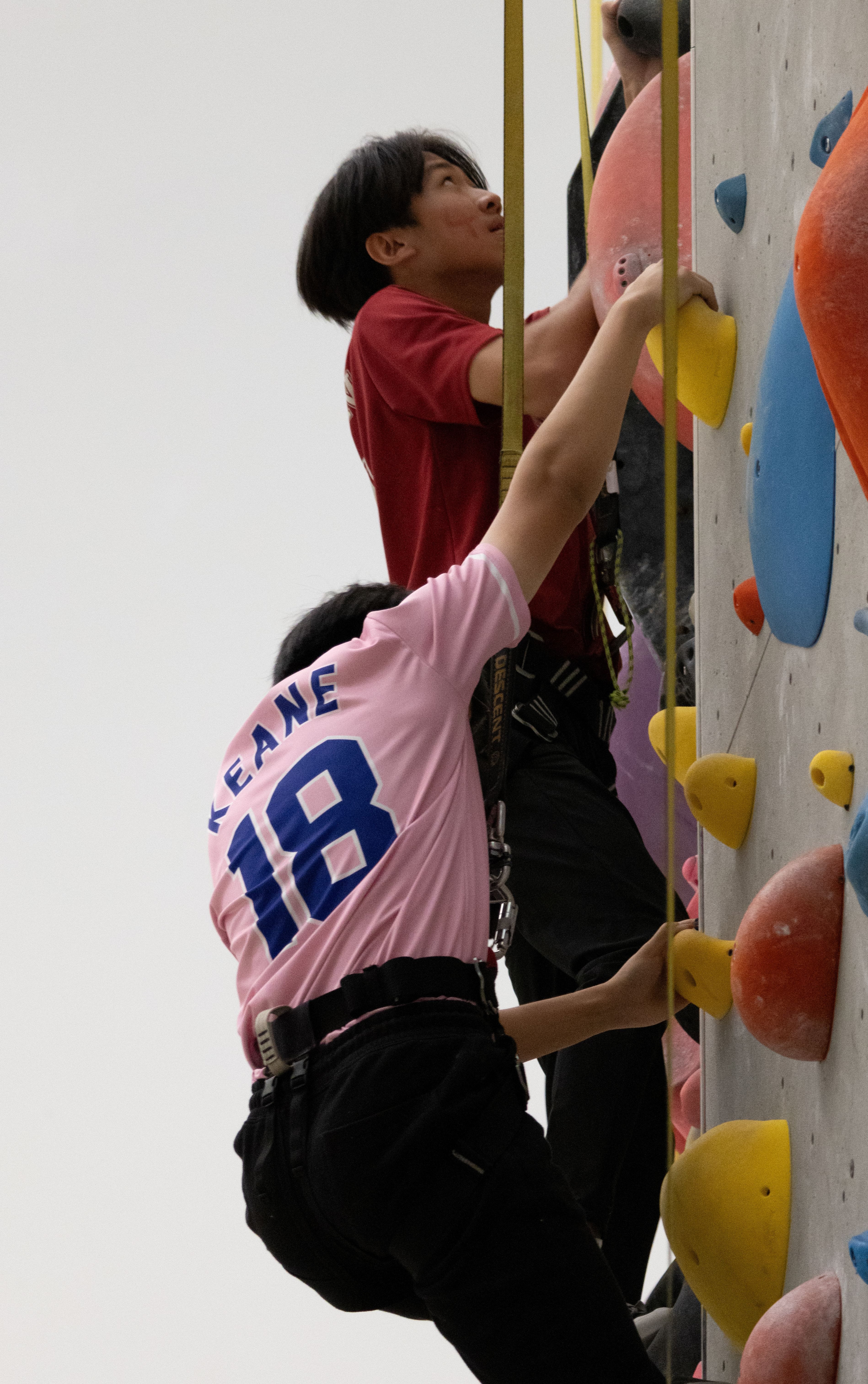 Two climbers on a rock wall with ropes; one wears a pink jersey reading KEANE 18 reaching upward.
