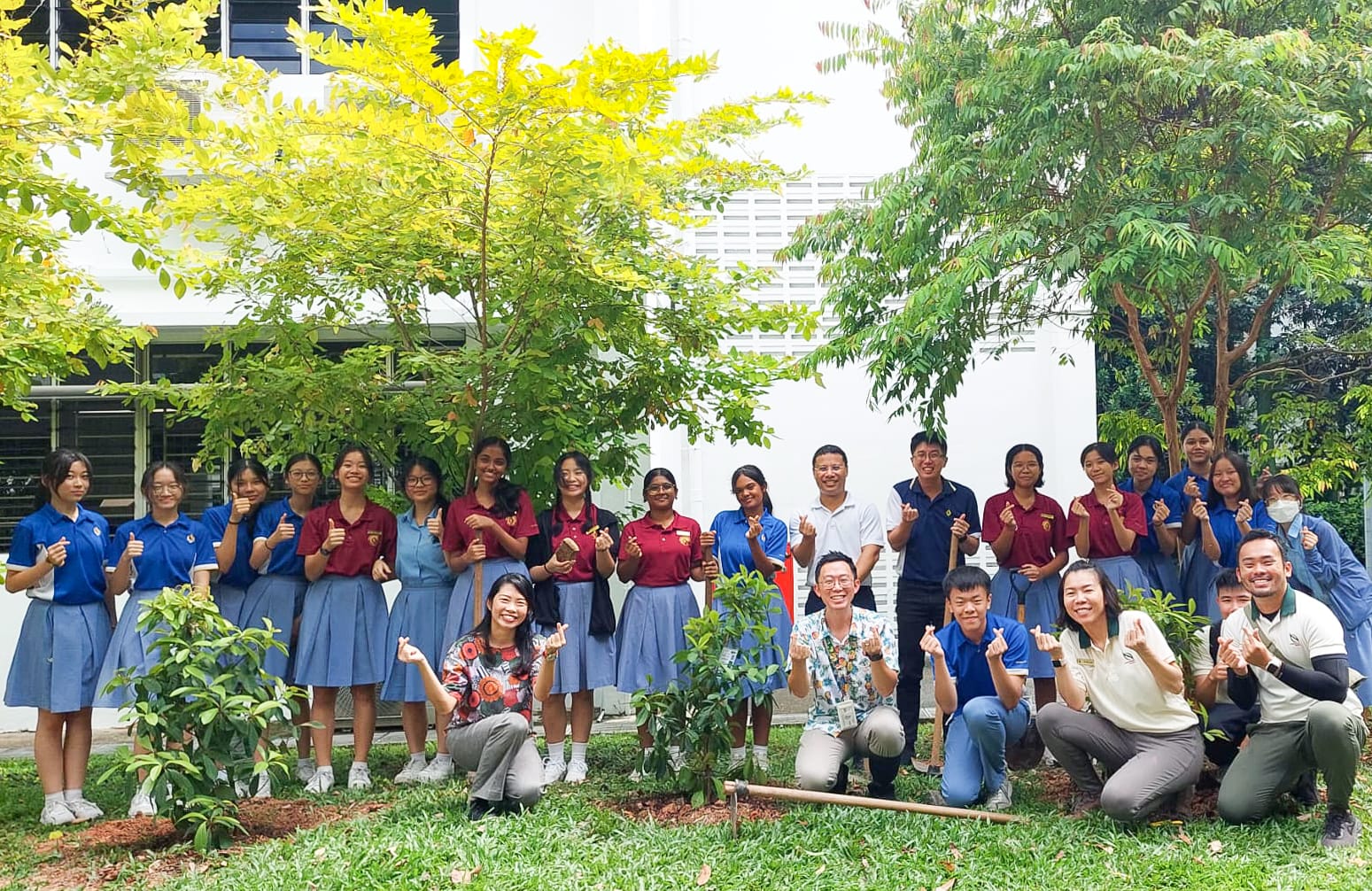 A large group of students in blue and maroon uniforms pose outdoors near trees, giving thumbs up and finger hearts.