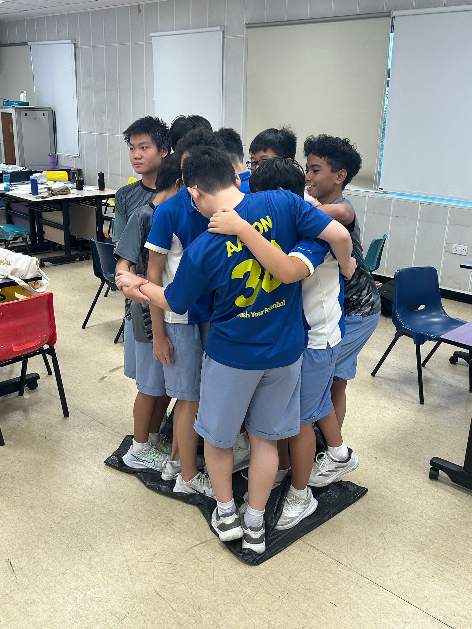 Group of students stand close together on a black mat in a classroom, arms around each other.