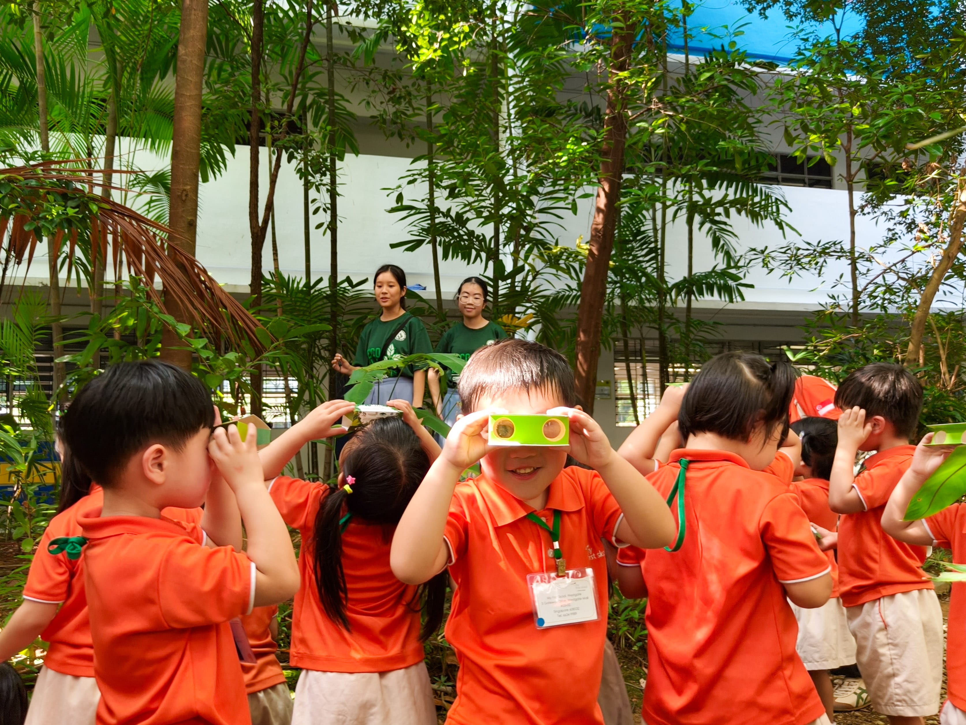 Young children in orange uniforms hold green cardboard binoculars to their eyes outdoors among trees.