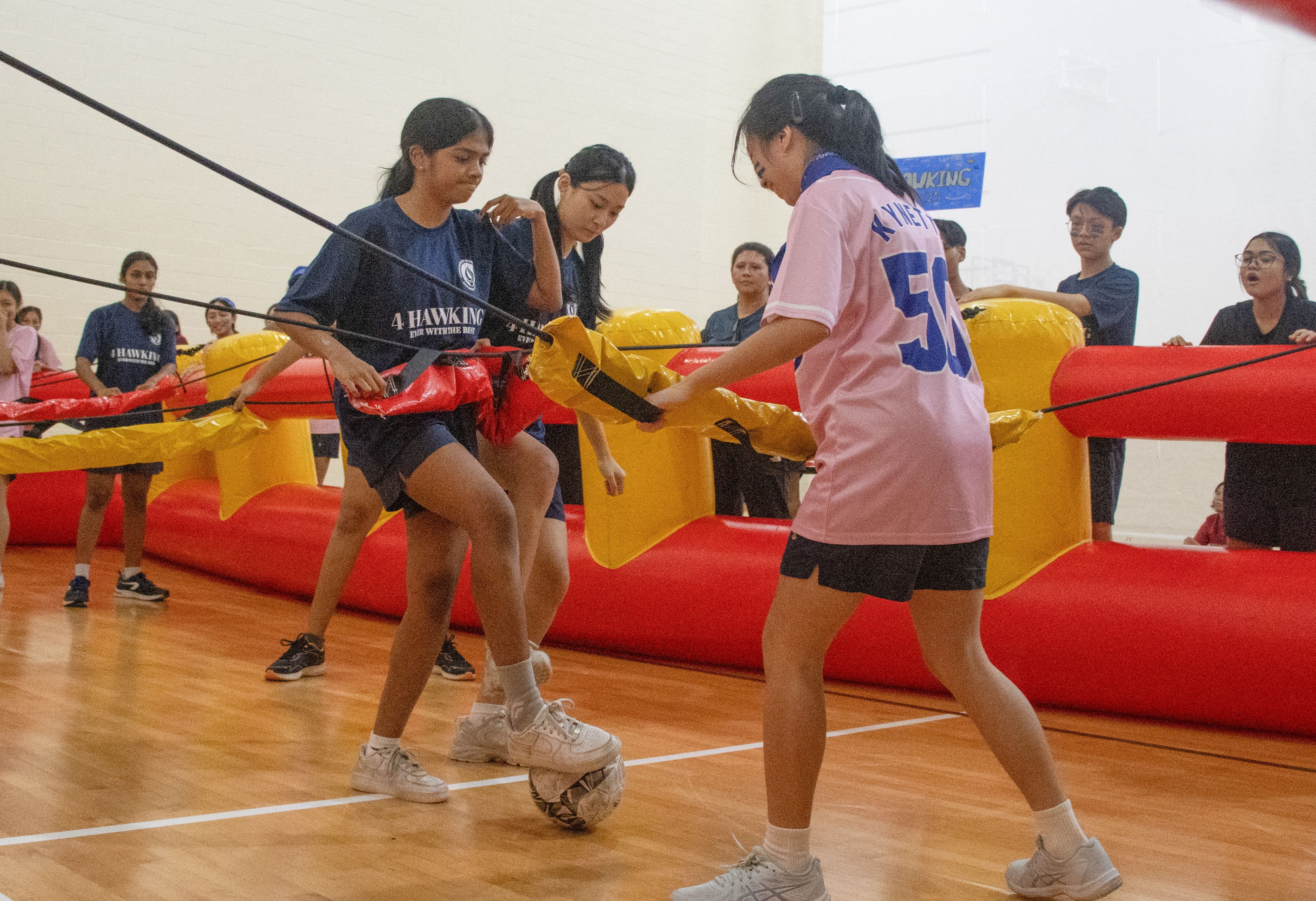 Two girls play bubble soccer in an inflatable arena while others watch, with a ball at their feet.