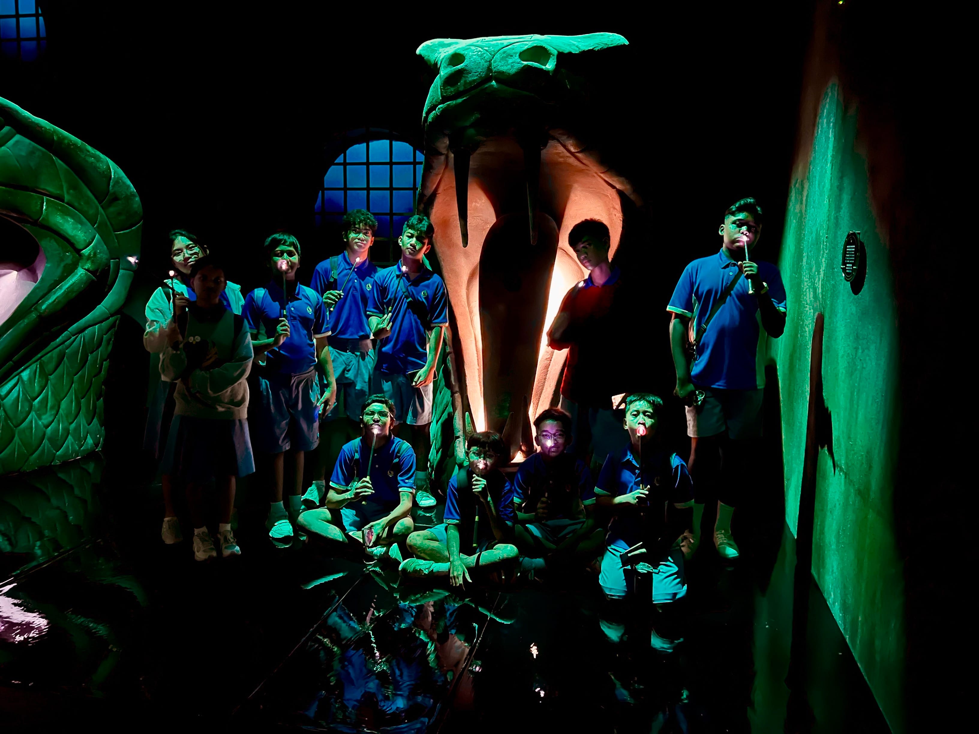 Students in blue uniforms pose inside a dark themed exhibit featuring a large green serpent and glowing creature skull.