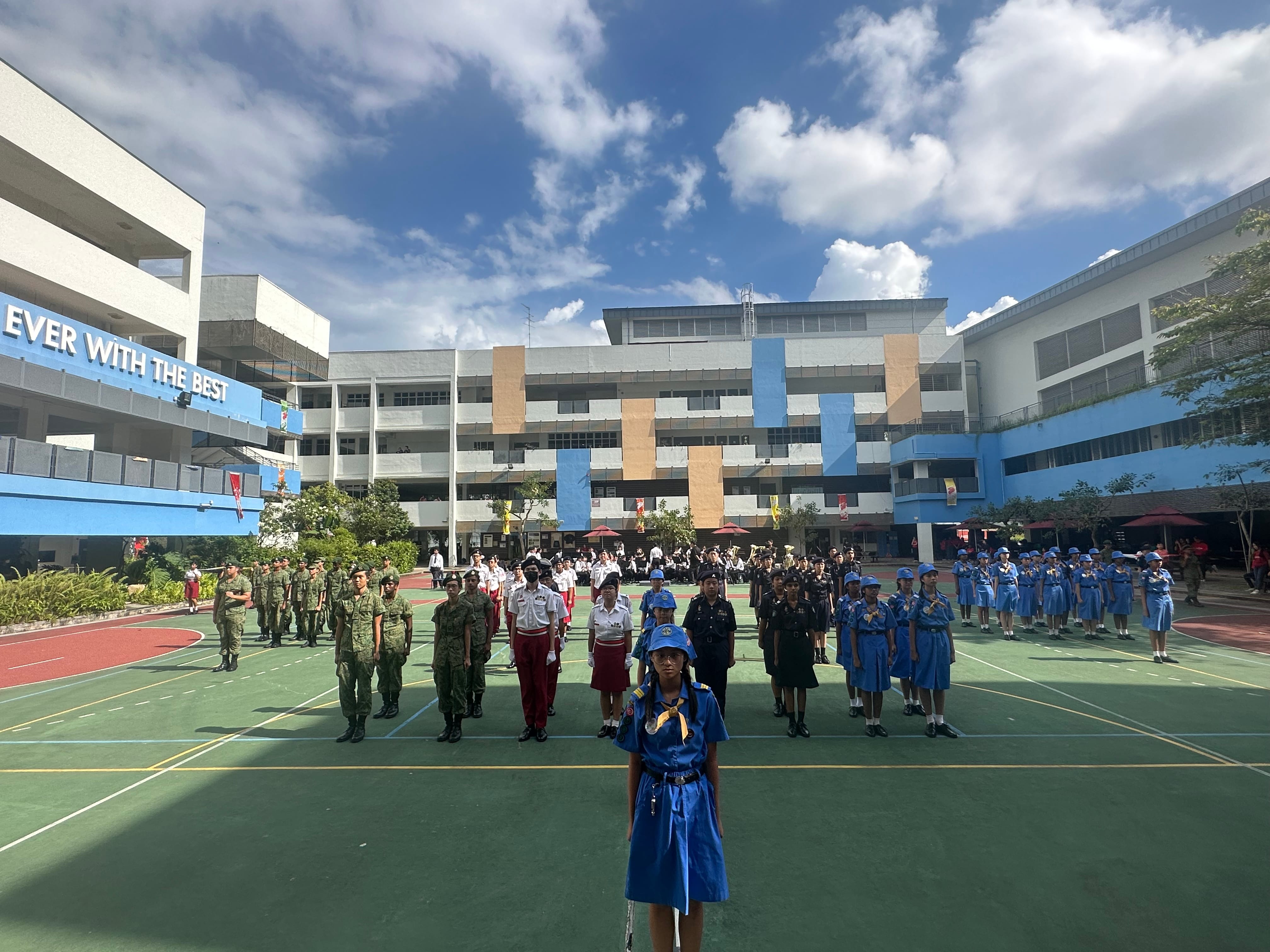 Students in various uniforms stand in formation on a school courtyard during an outdoor parade or assembly.