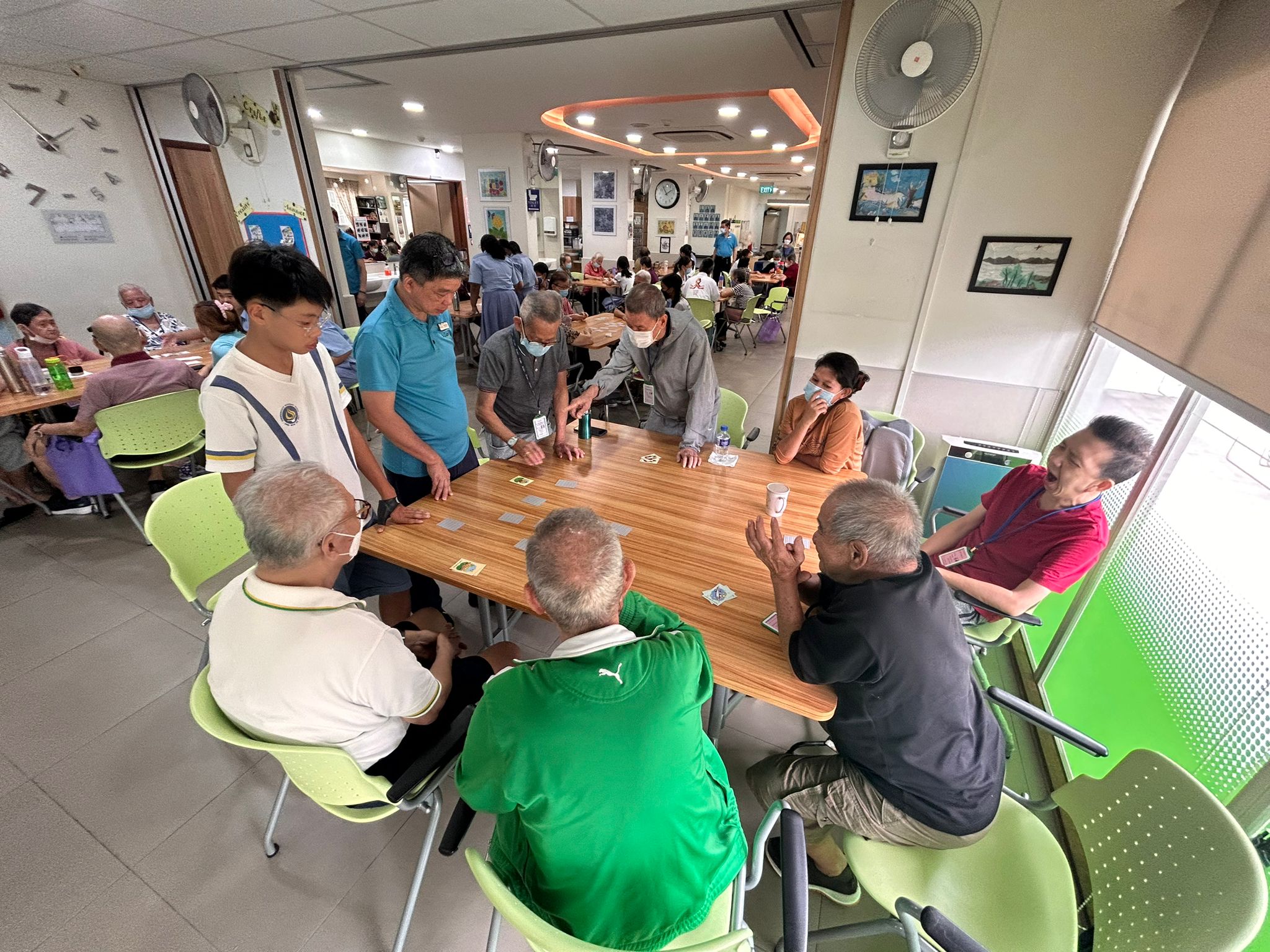 Several elderly adults play a card game at a wooden table with green chairs in a busy indoor community center.