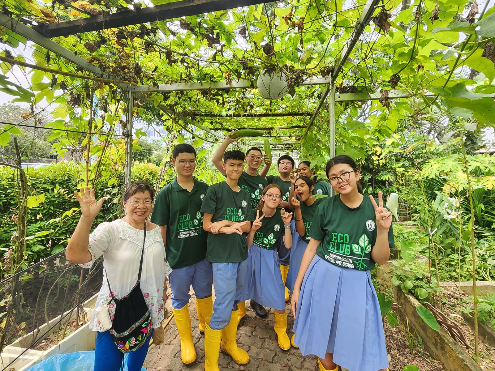 Students in green Eco Club shirts and yellow boots pose with an adult under a vine-covered pergola in a garden.