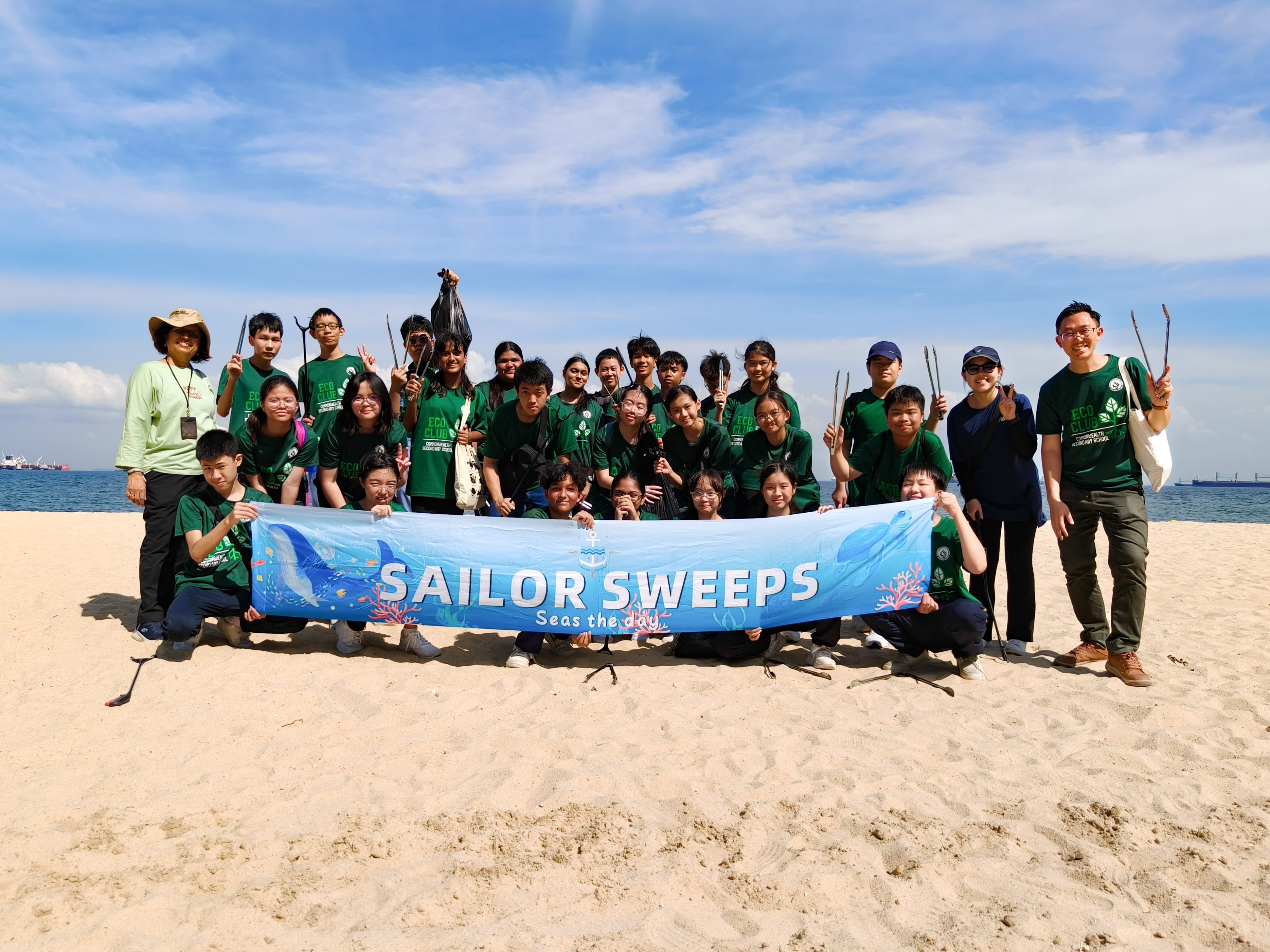 A group of students in green shirts pose on a beach holding a blue banner reading Sailor Sweeps Seas the Day.