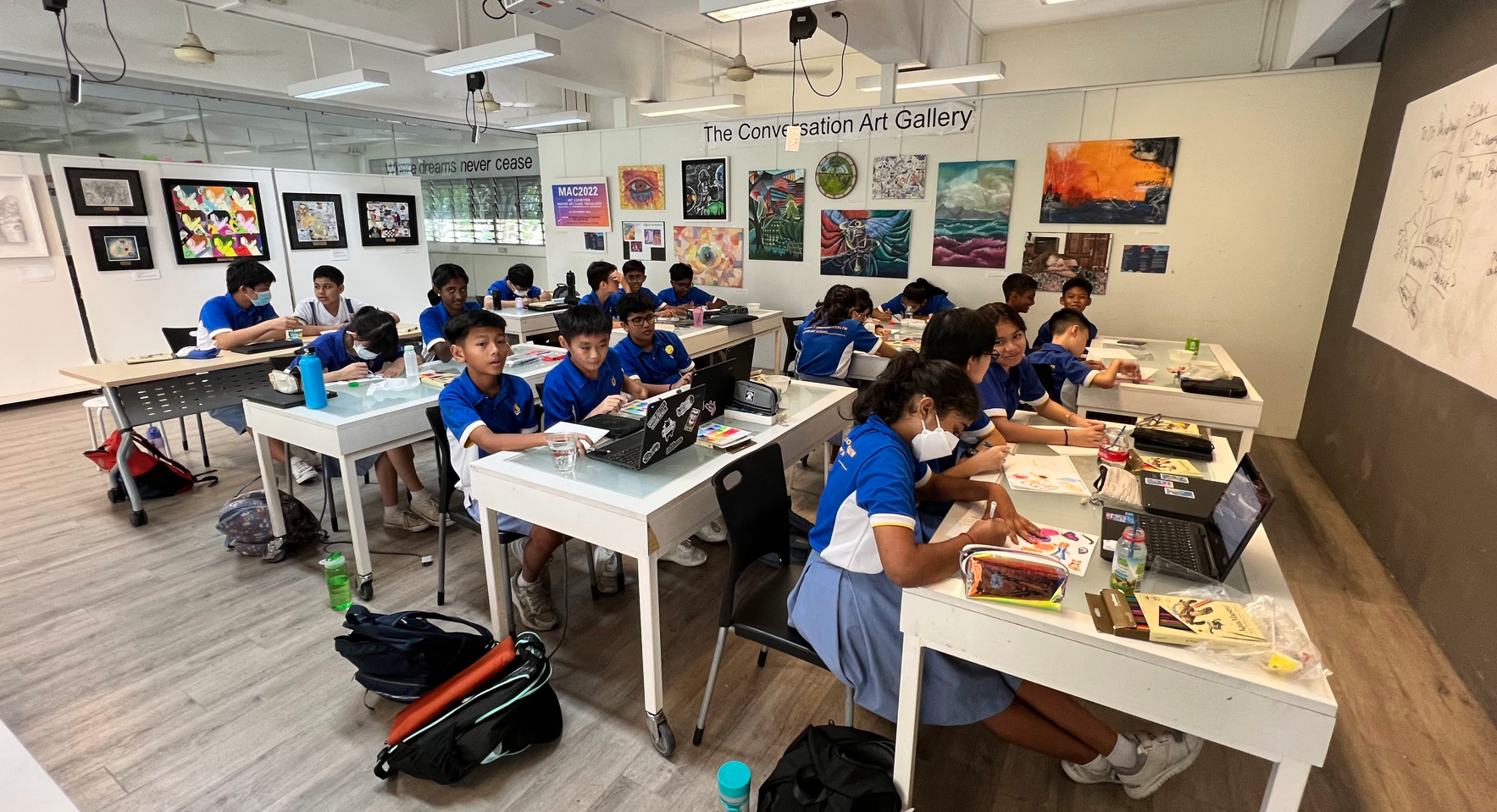 Students in blue uniforms sit at white desks in The Conversation Art Gallery classroom with colorful artwork on the walls.
