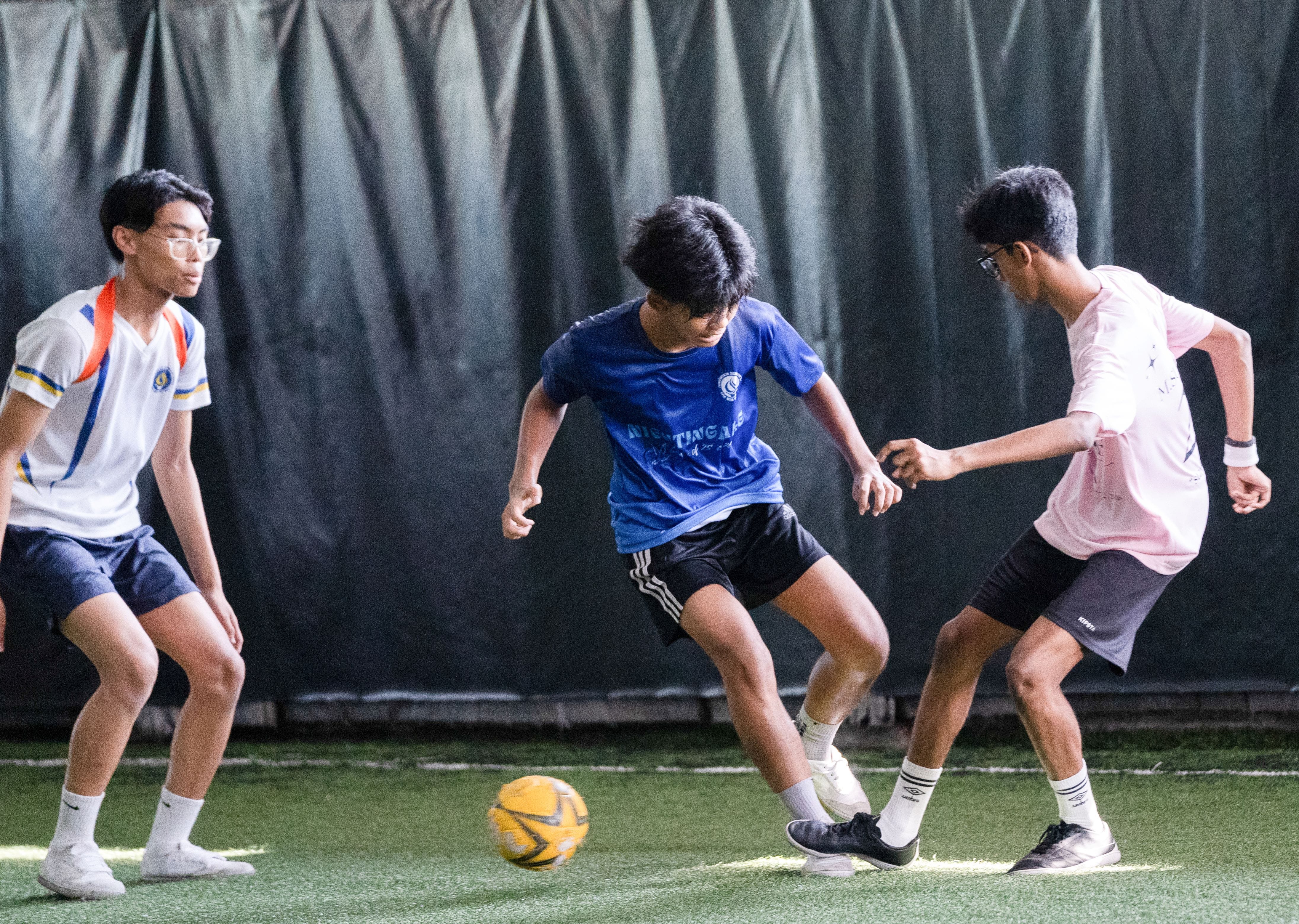 Three youths playing indoor soccer on turf, one dribbling a yellow ball between two defenders.