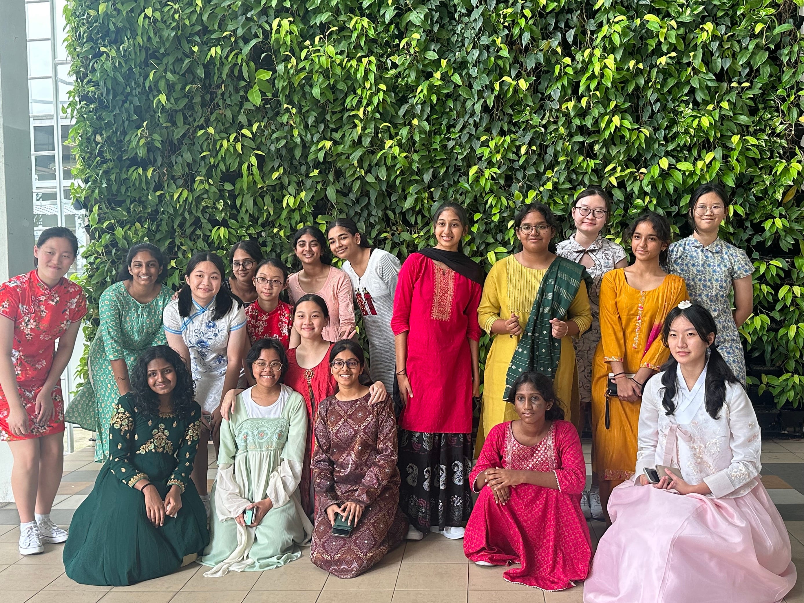 A group of young women in colorful traditional Asian and South Asian attire posing in front of a green living plant wall.
