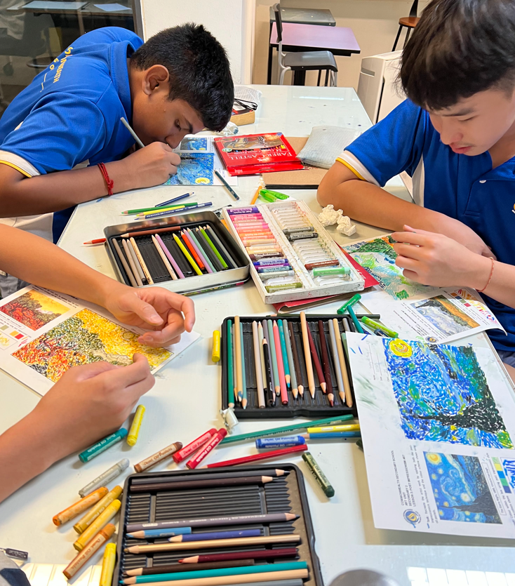 Three students in blue uniforms draw at a table covered with colored pencils, pastels, and art reference sheets.