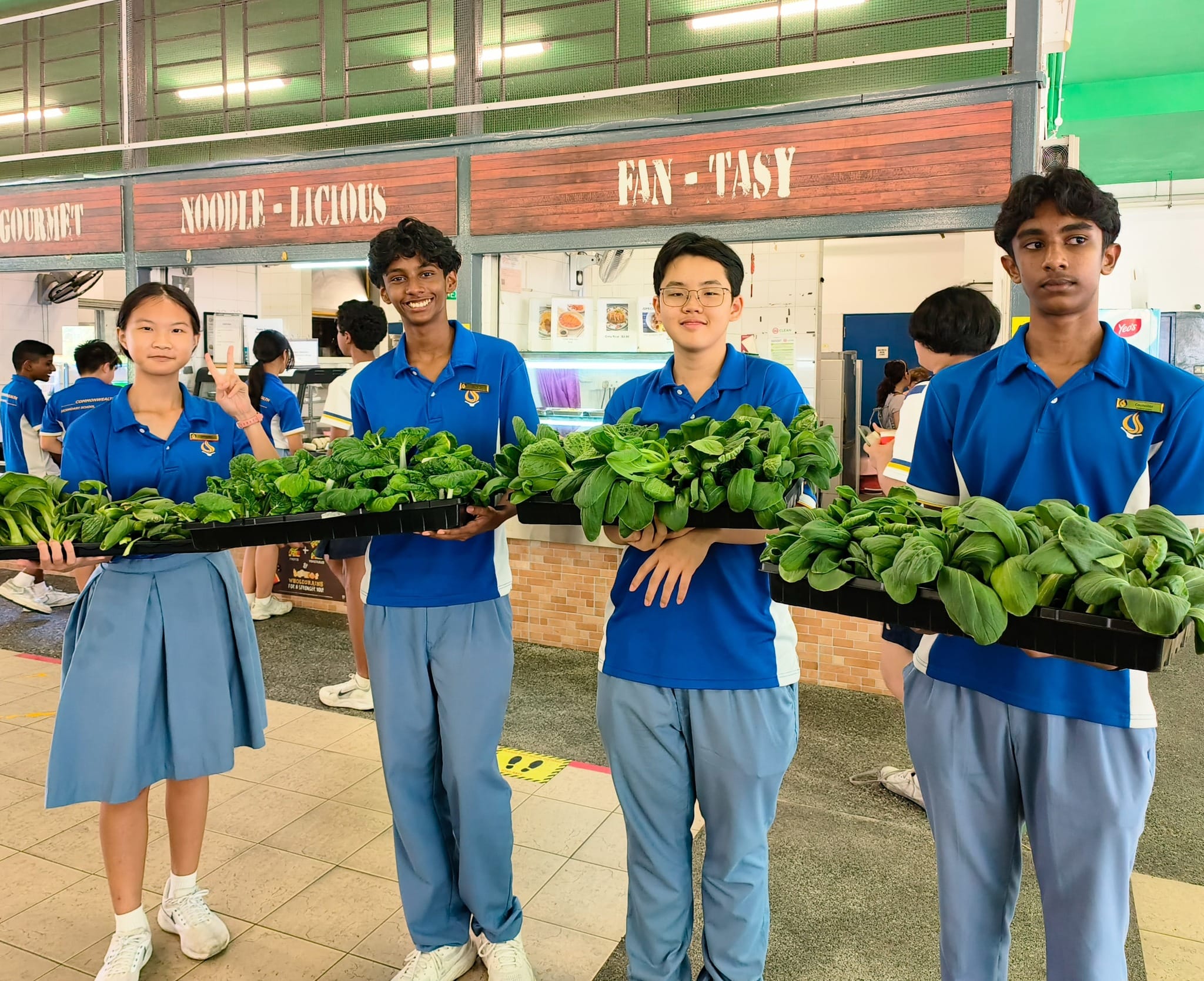 Students hold trays of leafy greens in a school canteen; one flashes a peace sign.