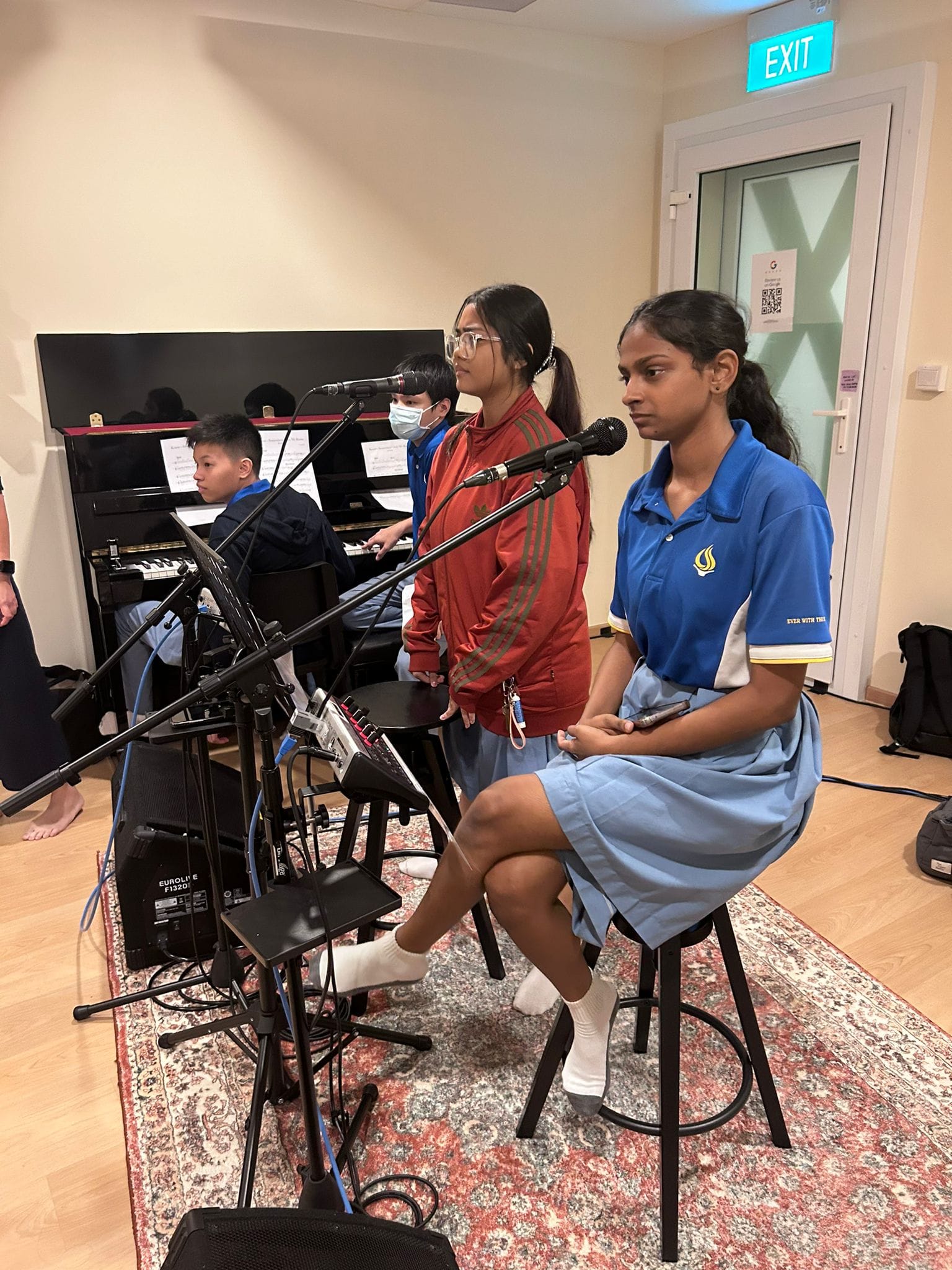 Two girls at microphones in a rehearsal room while two boys play upright piano; EXIT sign over a door.