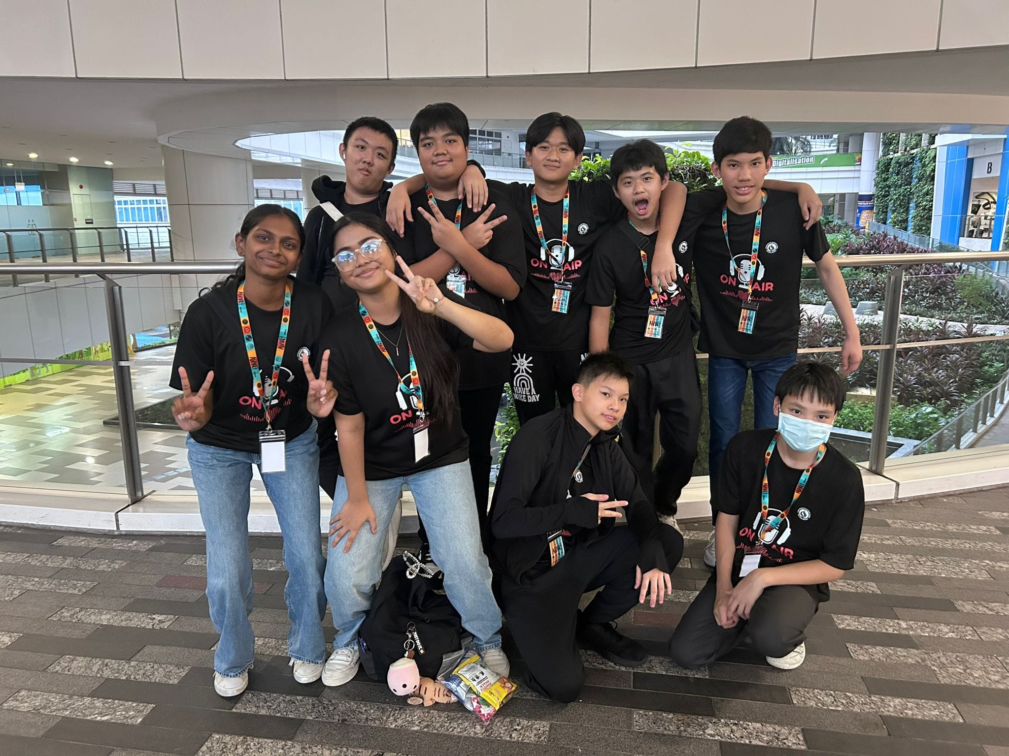 Nine teens in black T-shirts and lanyards pose indoors; some flash peace signs, one wears a mask.