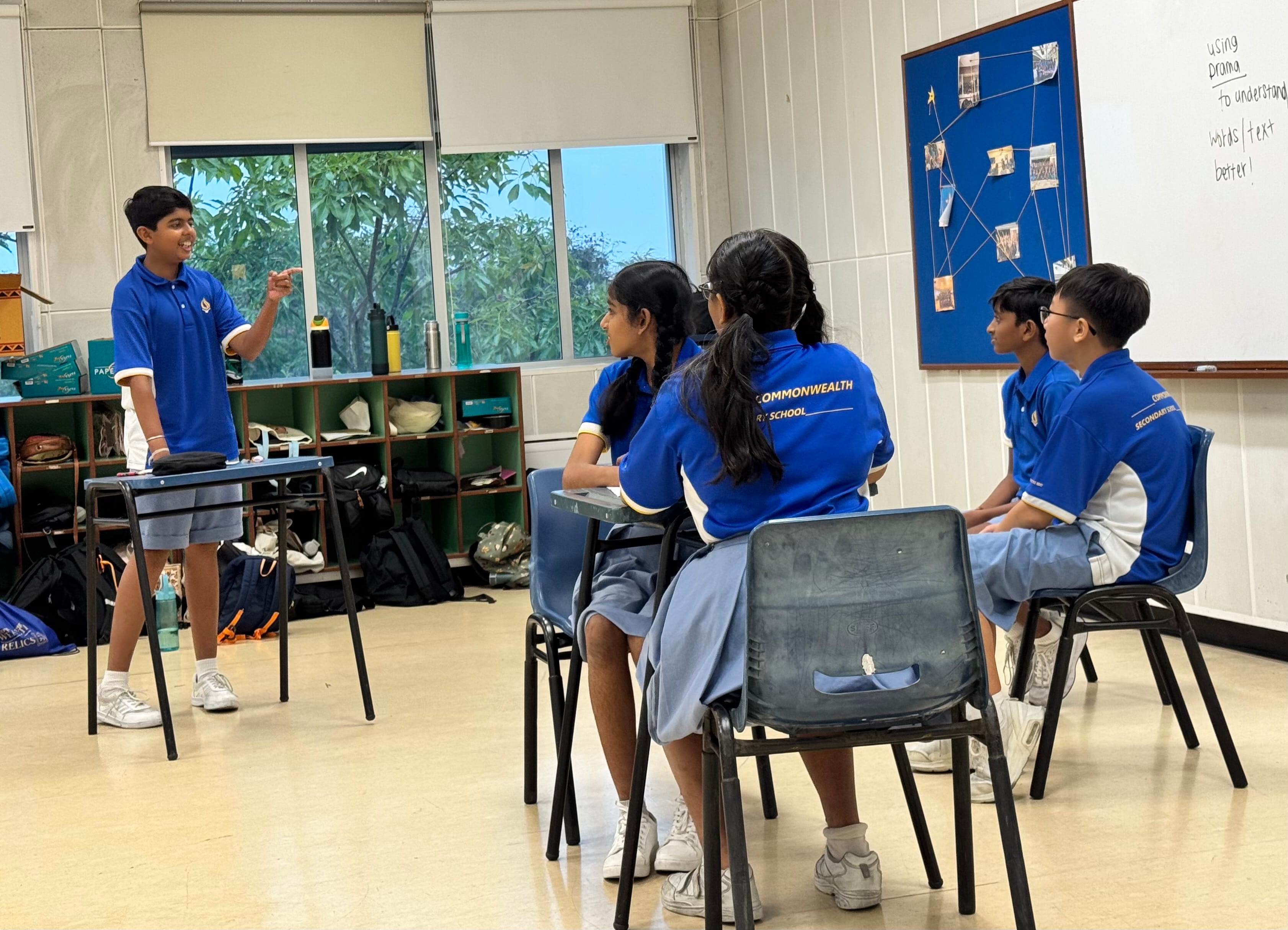 A student in a blue uniform stands and speaks to four seated classmates in a school classroom.