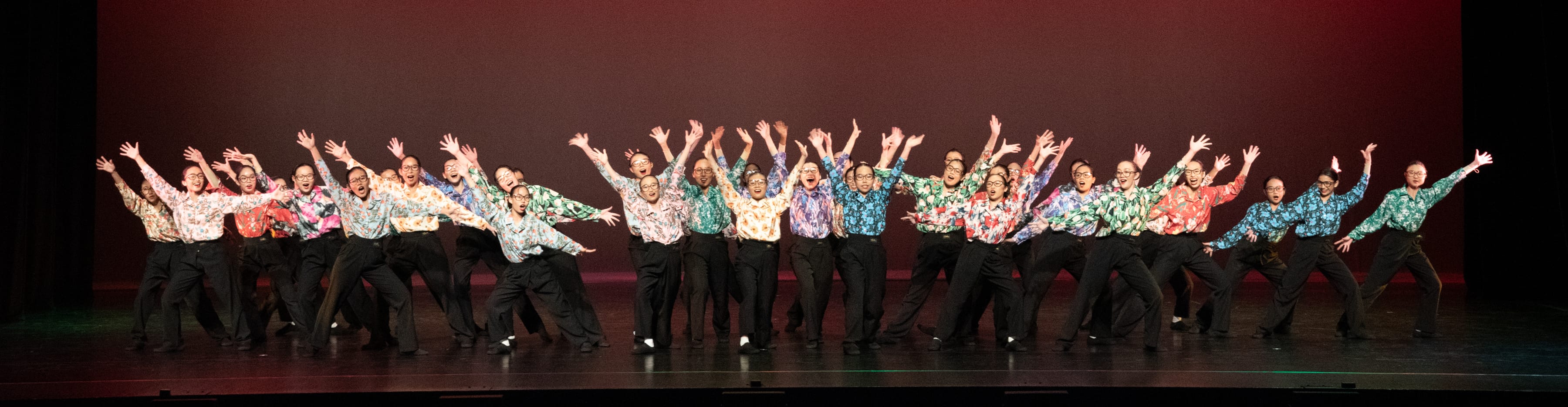 Large group of performers in colorful floral shirts and black pants dancing on stage with arms raised.