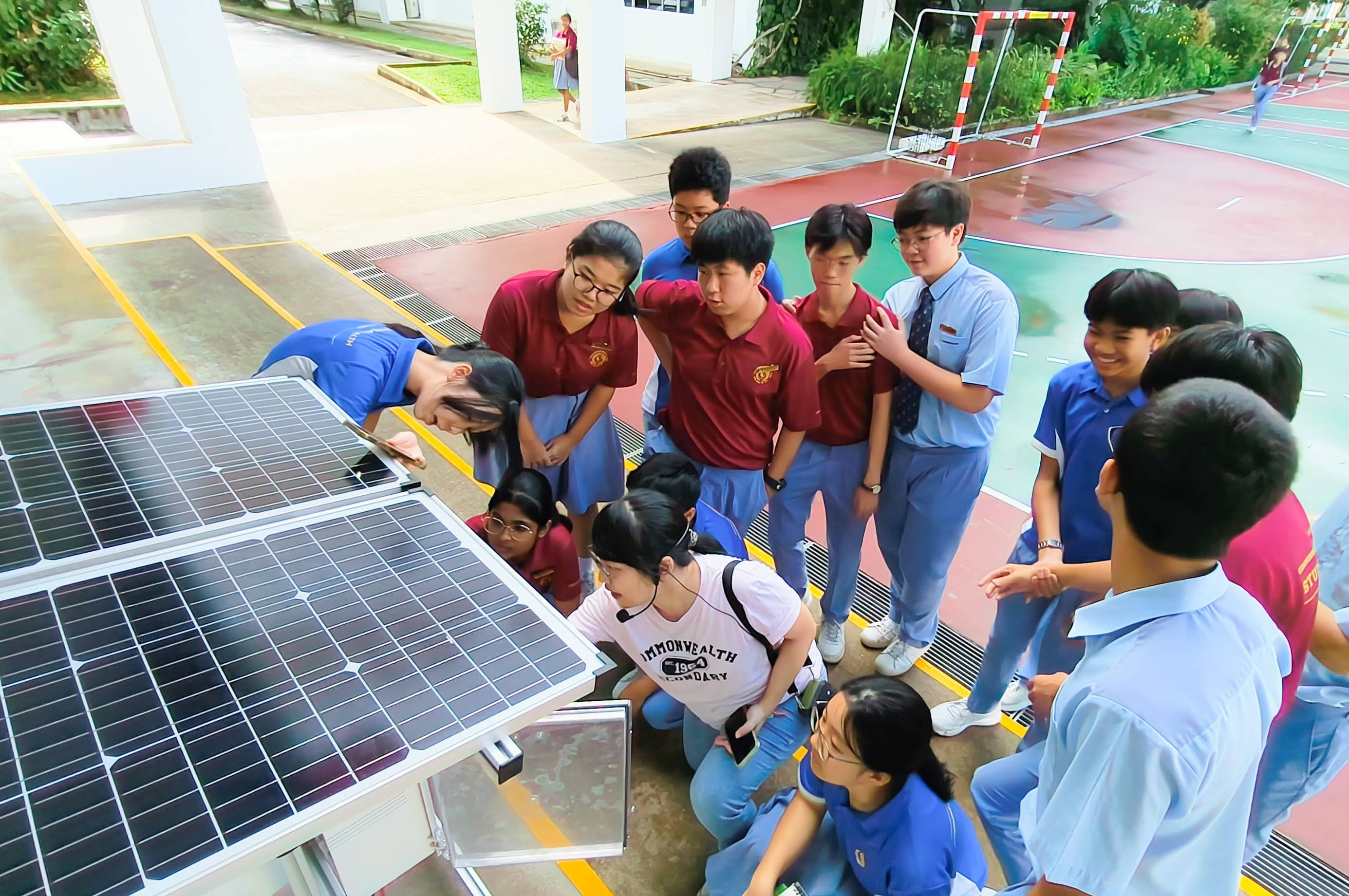 A group of school students in uniforms gather closely around two solar panels outdoors on a school campus.