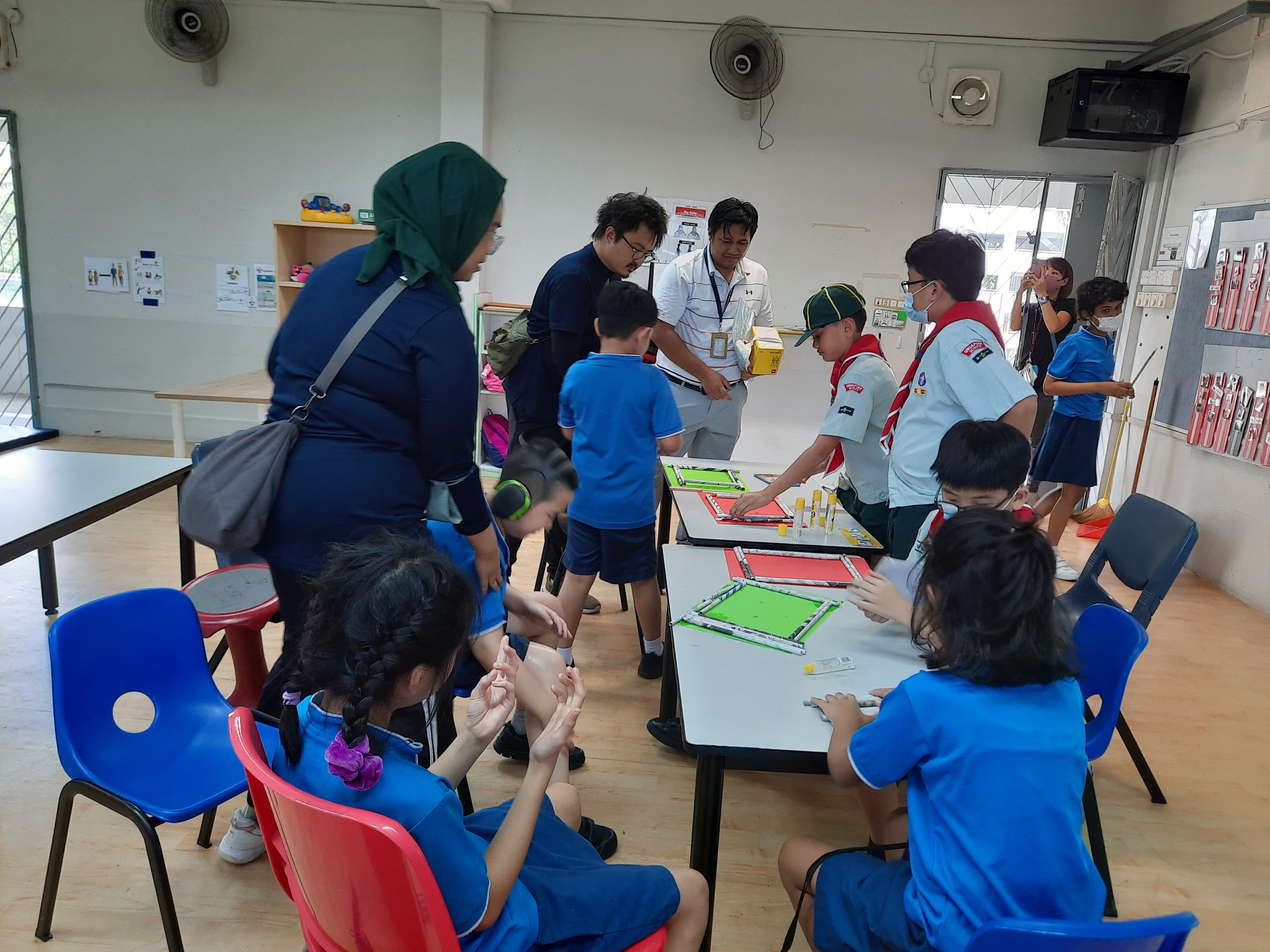 Children in blue uniforms and scouts gather around tables with green board game activity in a classroom.