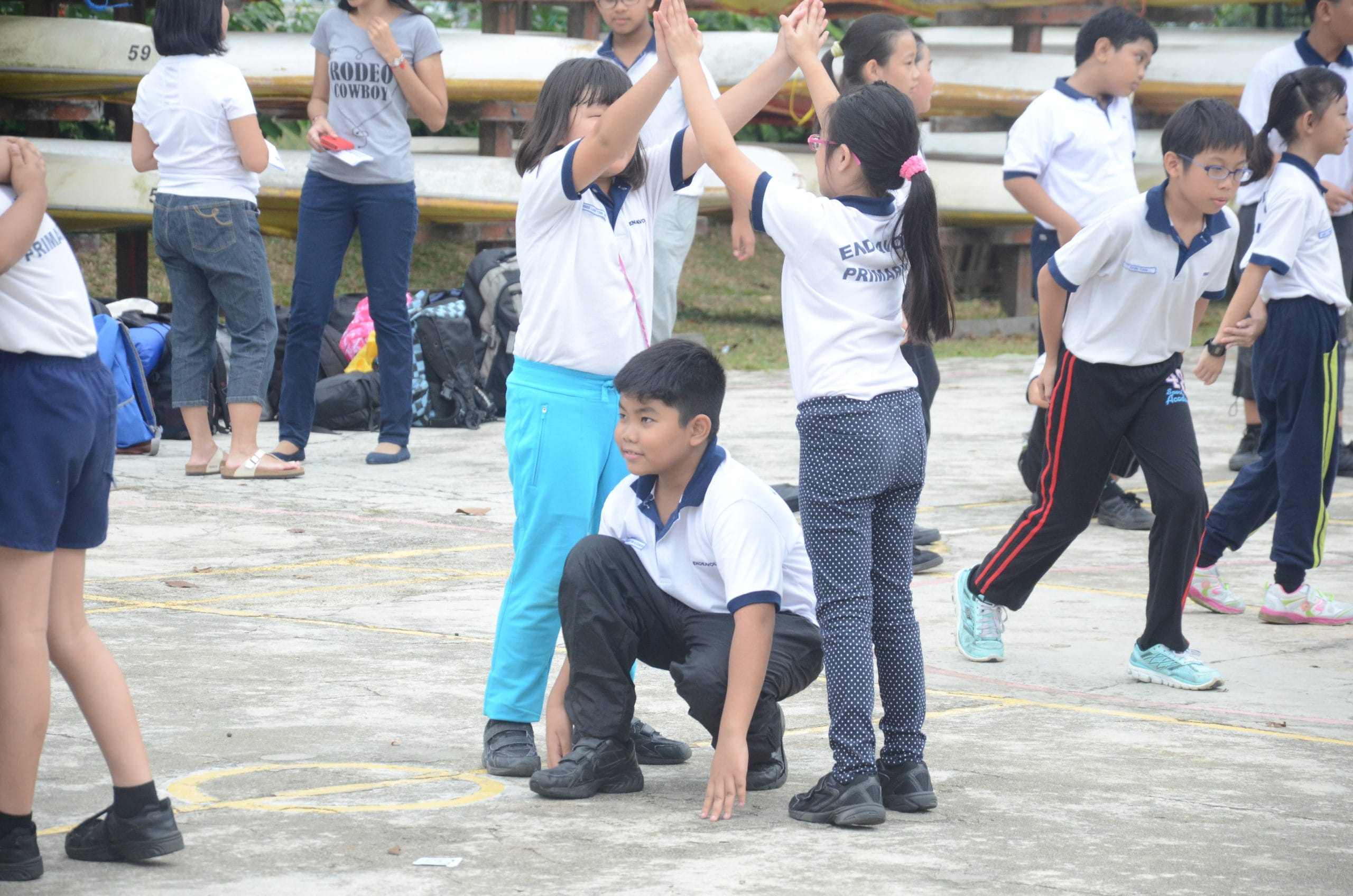 Primary school children in uniforms play outdoors; two girls high-five while a boy crouches between them.