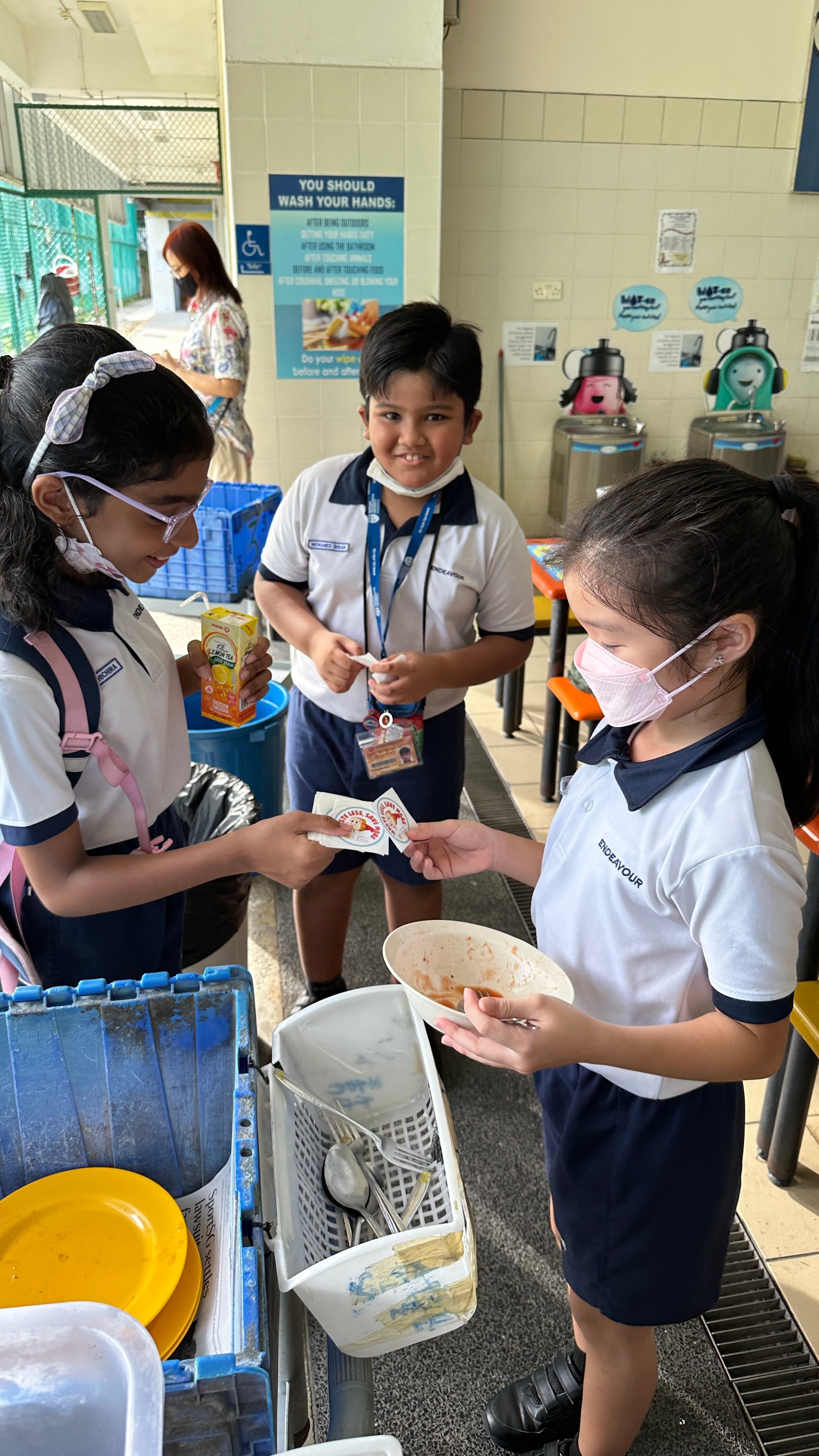 Three school children in uniforms and masks return dirty dishes and utensils at a school canteen tray return station.