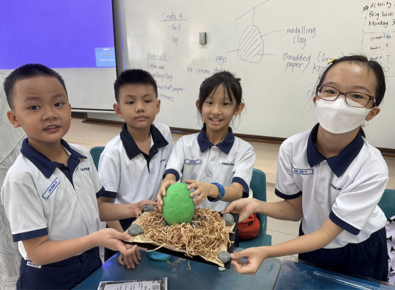 Four primary school students proudly holding up a group project featuring a green clay egg inside a nest made of shredded paper.