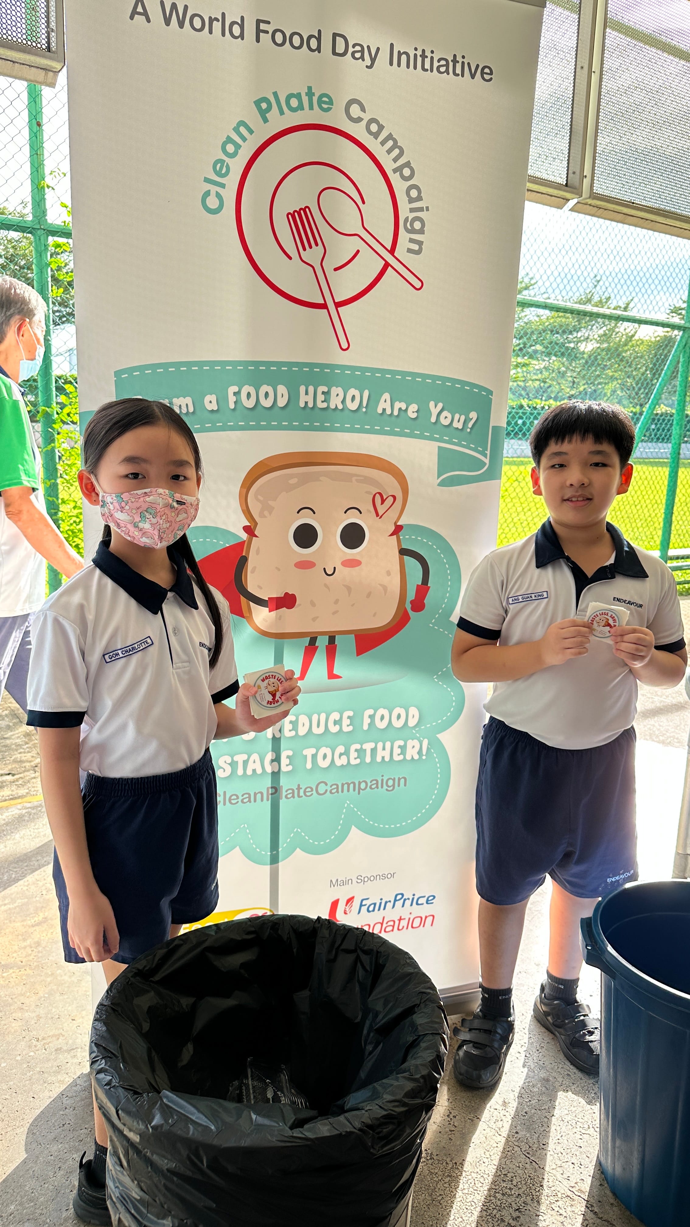 Two school children hold badges in front of a Clean Plate Campaign World Food Day banner outdoors.