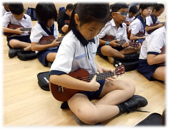 Several school children in uniforms sit on a wooden floor playing ukuleles during a music lesson.