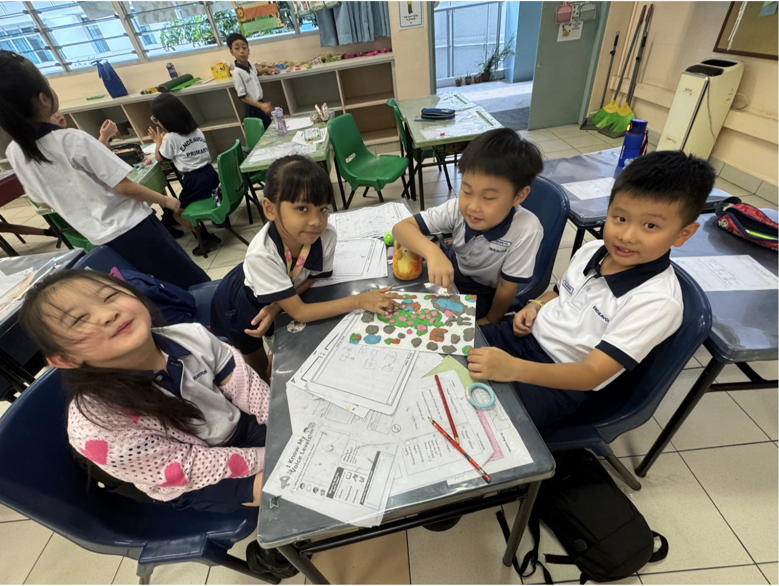 Four primary school students seated together at classroom desks, collaborating on a creative project using craft materials and worksheets.