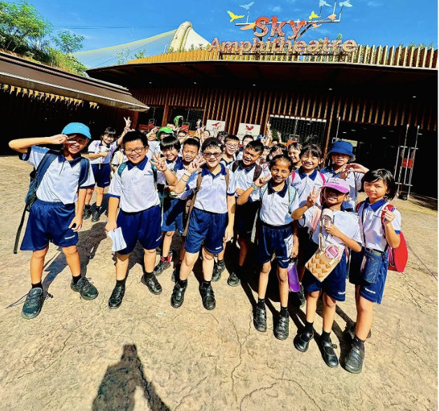 A group of uniformed schoolchildren posing cheerfully in front of the Sky Amphitheatre building outdoors.