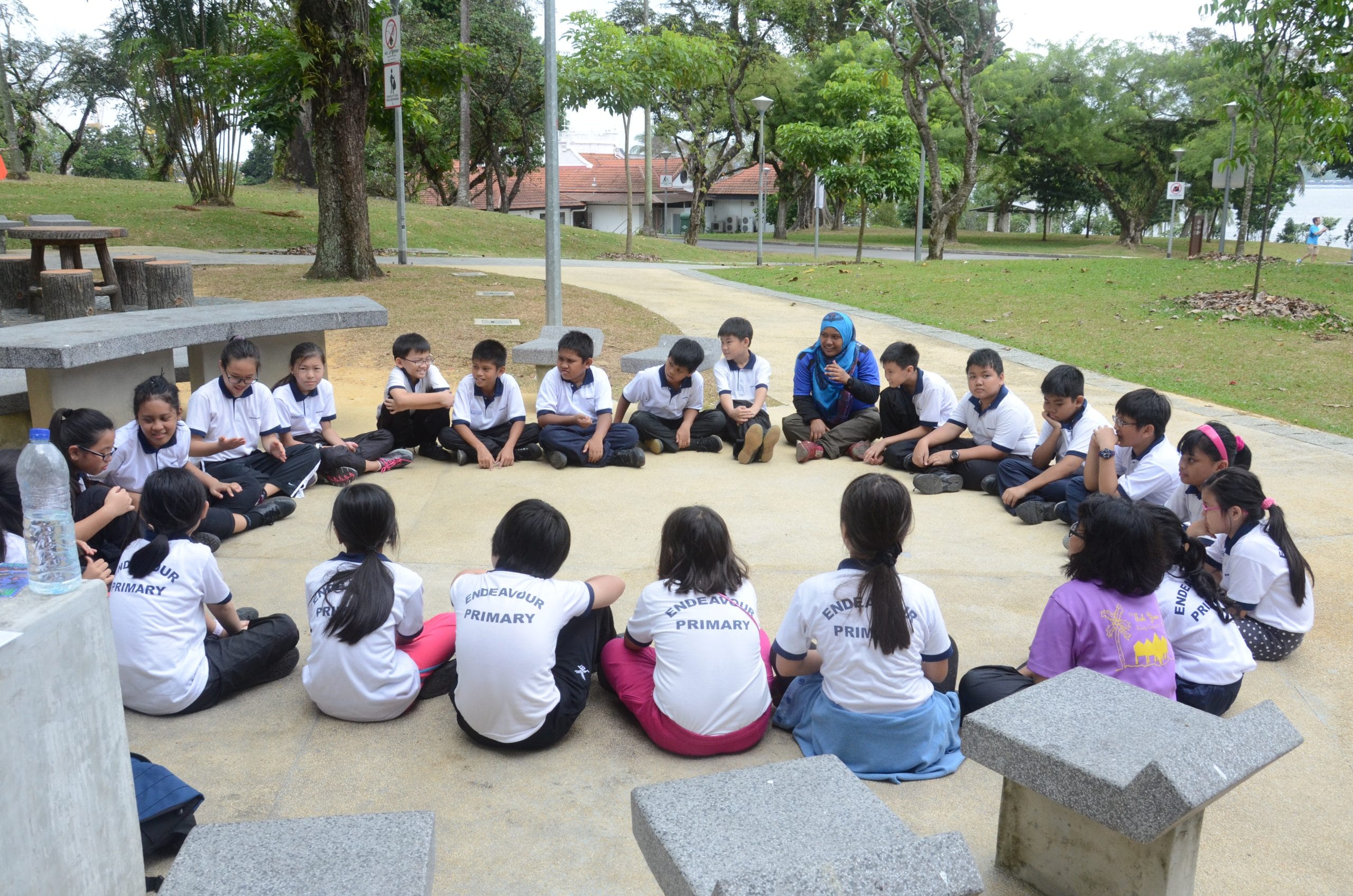 Endeavour Primary school children sit in a circle on the ground outdoors with a teacher in a park setting.