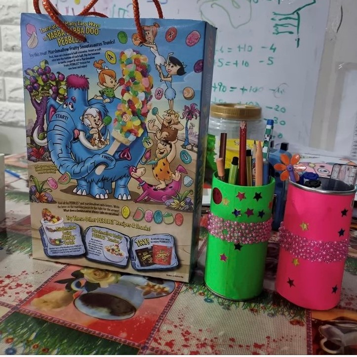Flintstones Pebbles cereal box beside two decorated pencil holders on a desk with math notes on the wall.