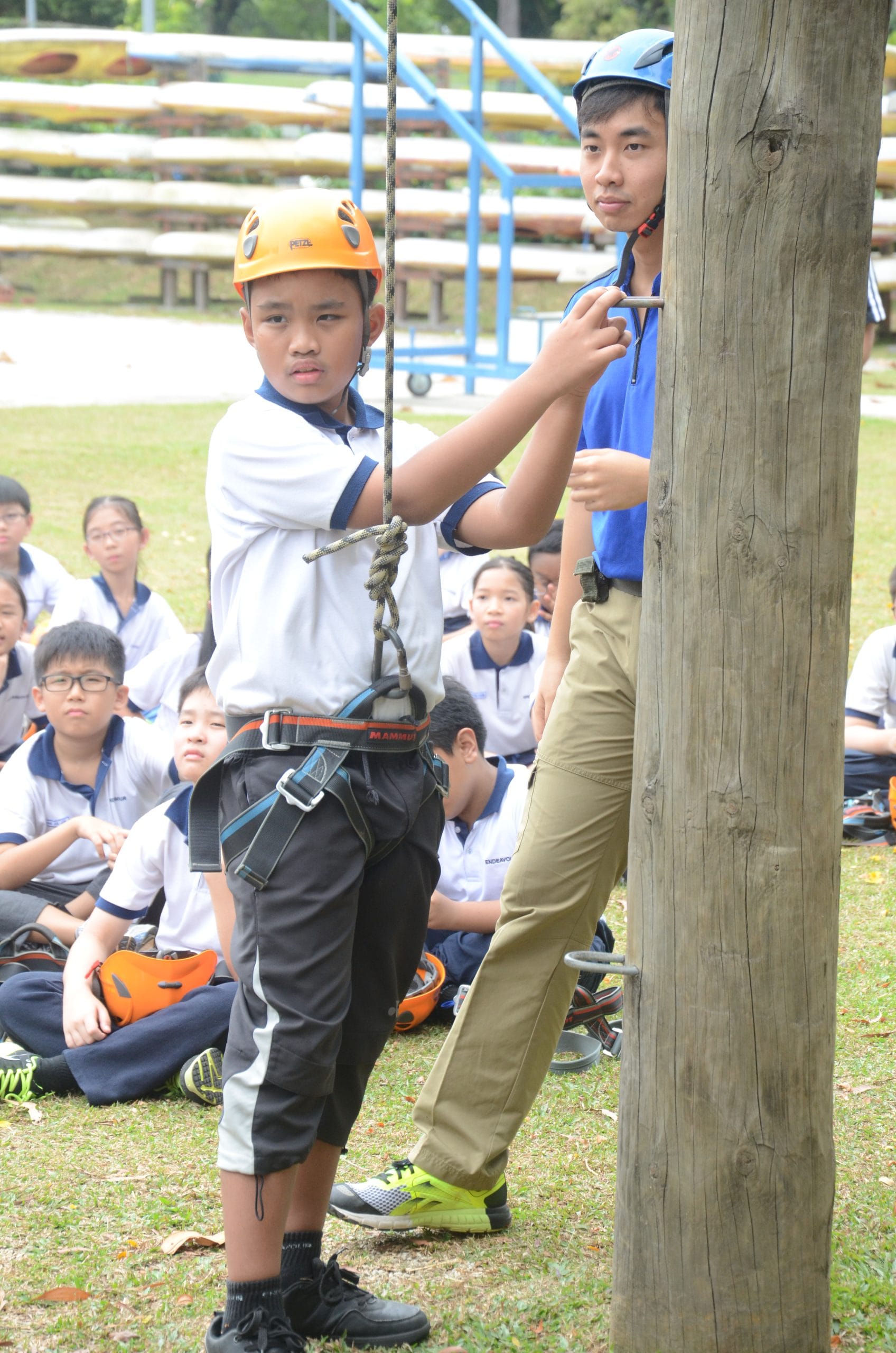 Two boys in helmets and harnesses practice rope climbing on a wooden pole while classmates watch seated on grass.