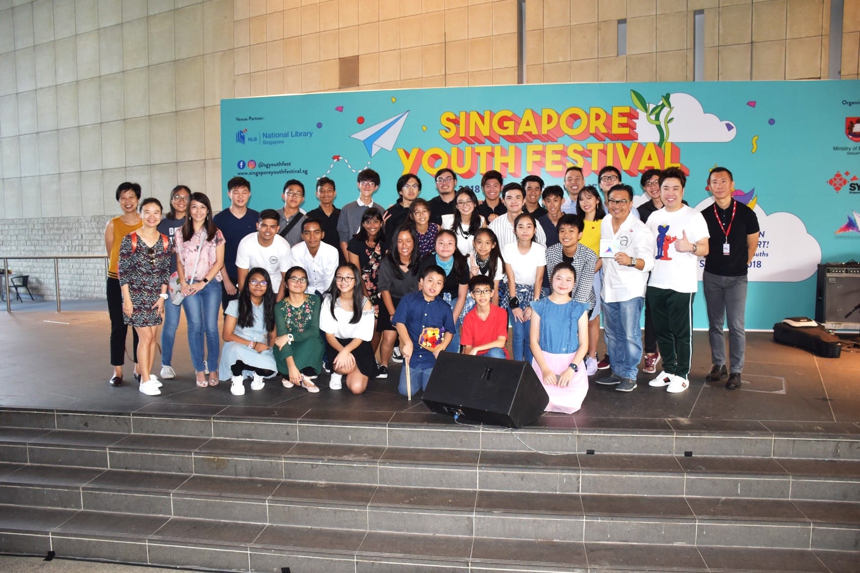A large group of young people posing on steps in front of a Singapore Youth Festival 2018 banner.
