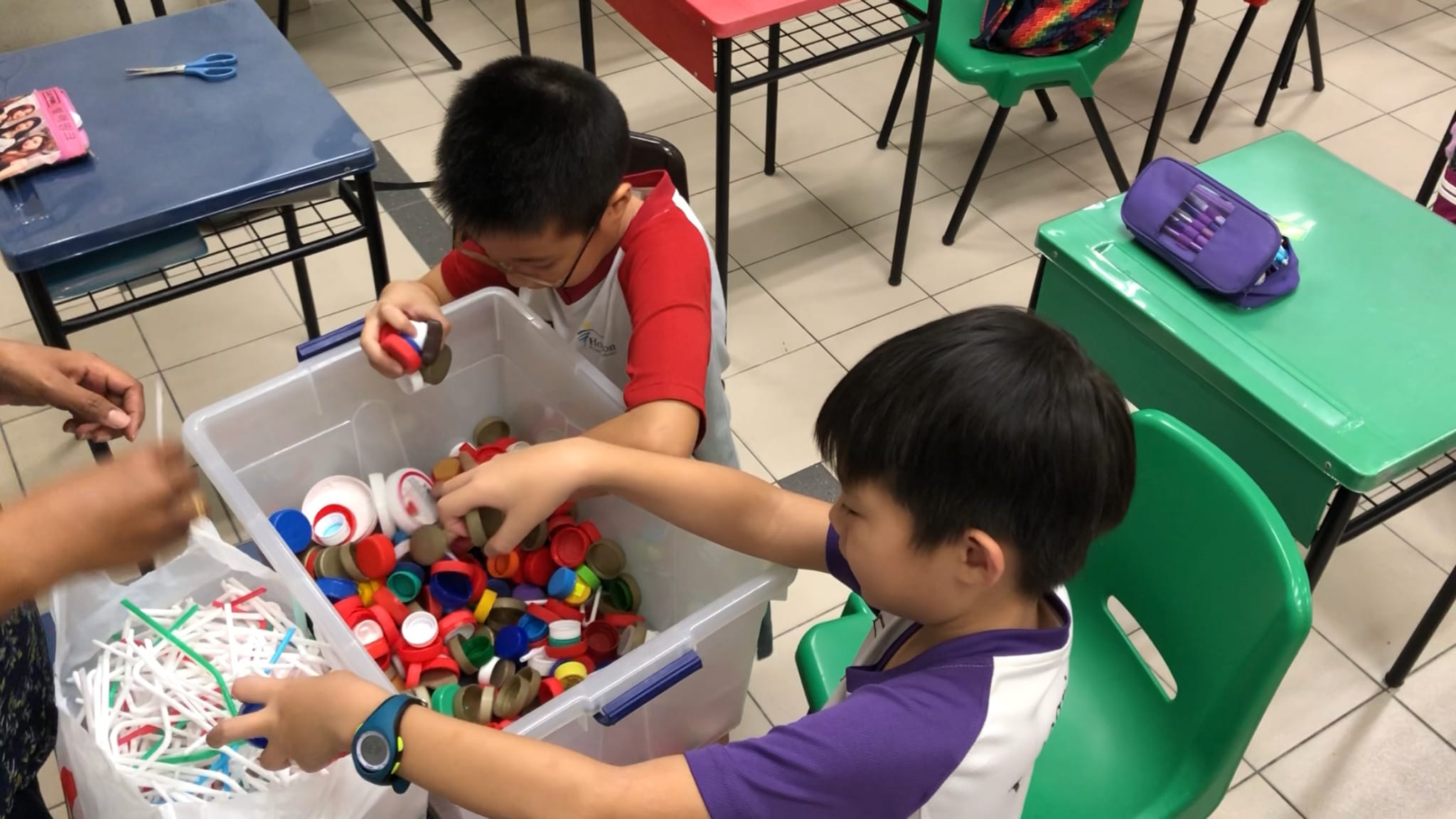 Two children sort colorful plastic bottle caps from a large bin in a classroom setting.