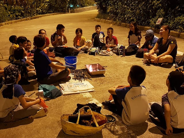Children and adults sit in a circle on pavement at night around a small fire in a metal tray.