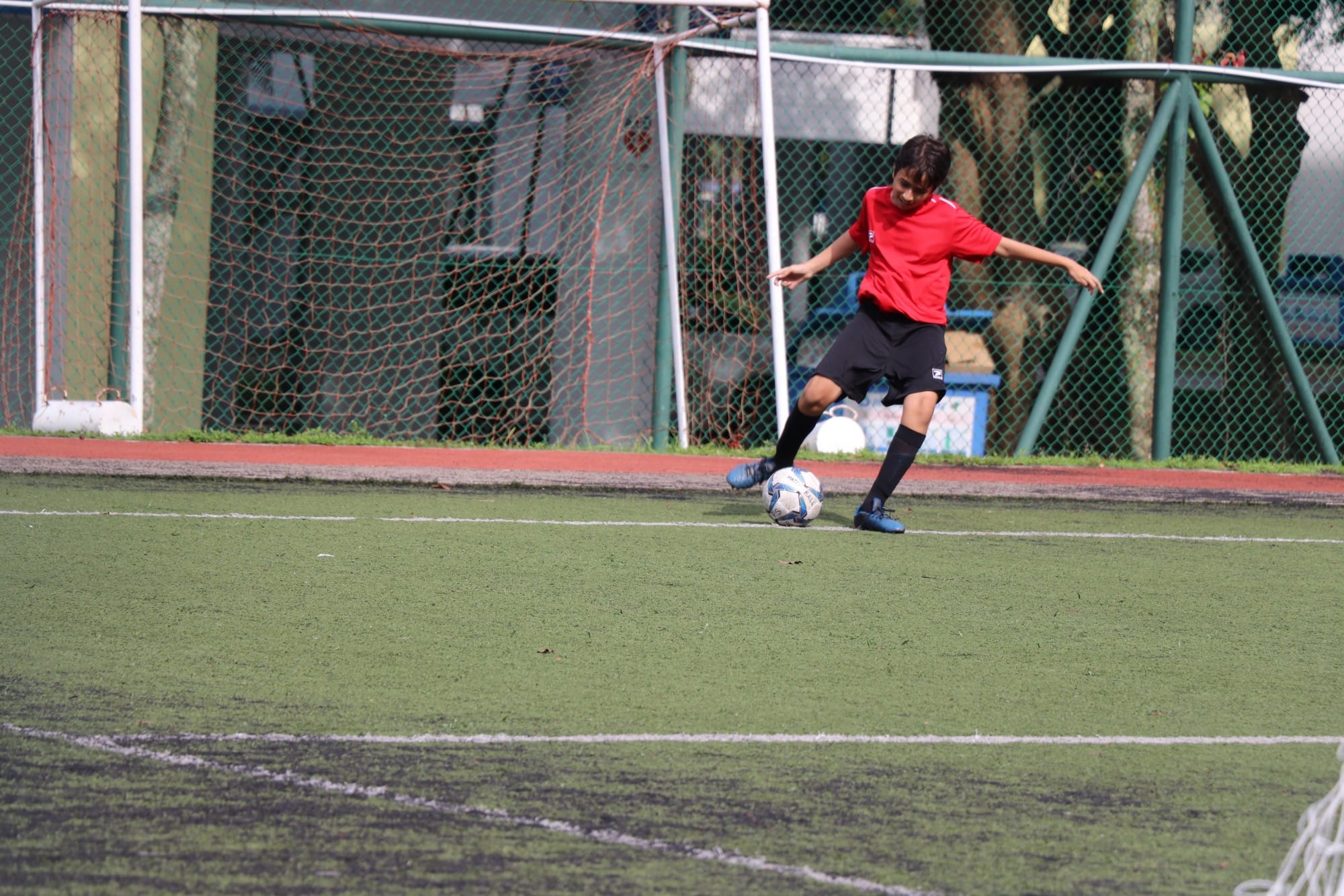 A young player in a red shirt and black shorts kicks a soccer ball on a green artificial turf field.