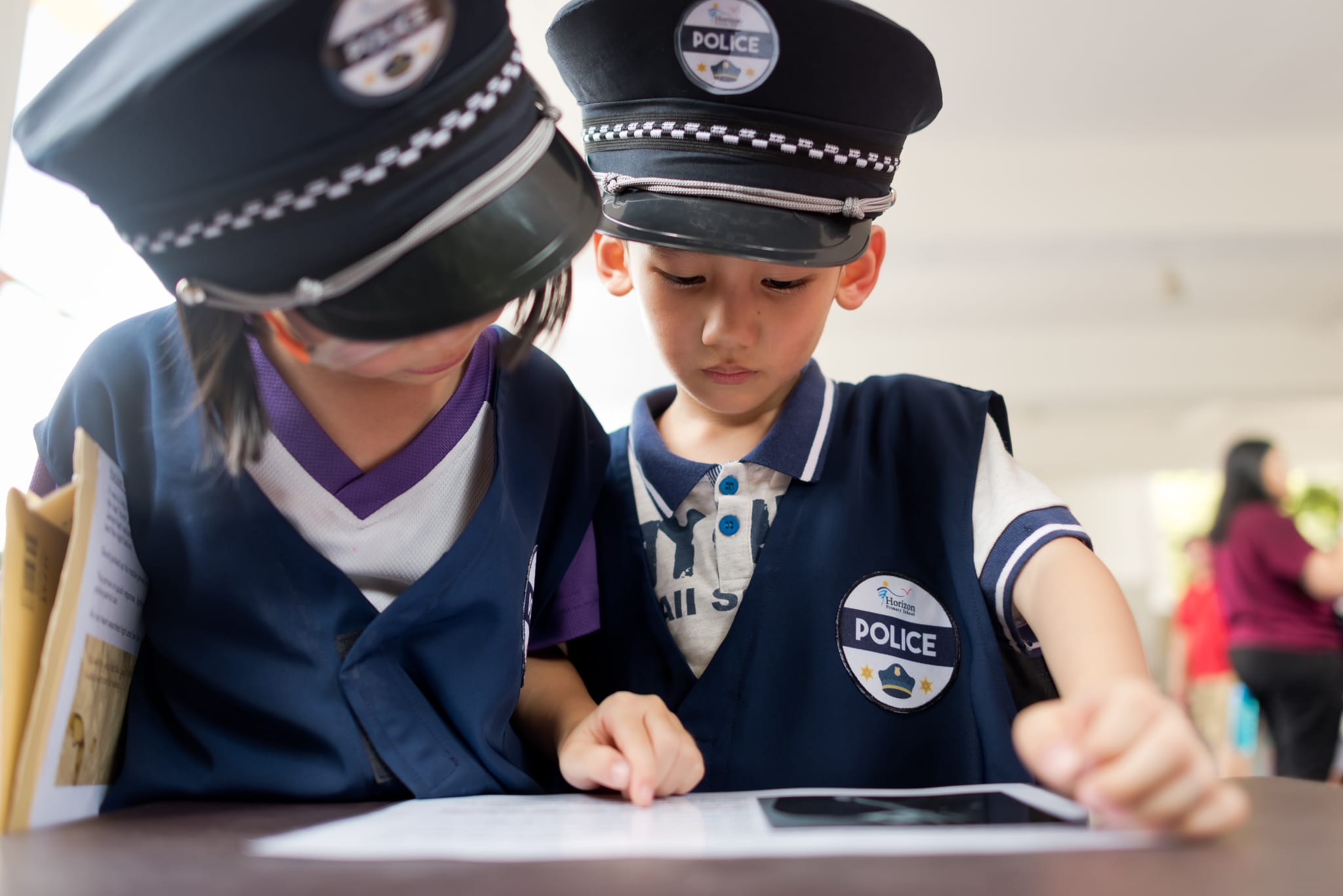 Two young children wearing toy police hats and vests look down at papers on a table.