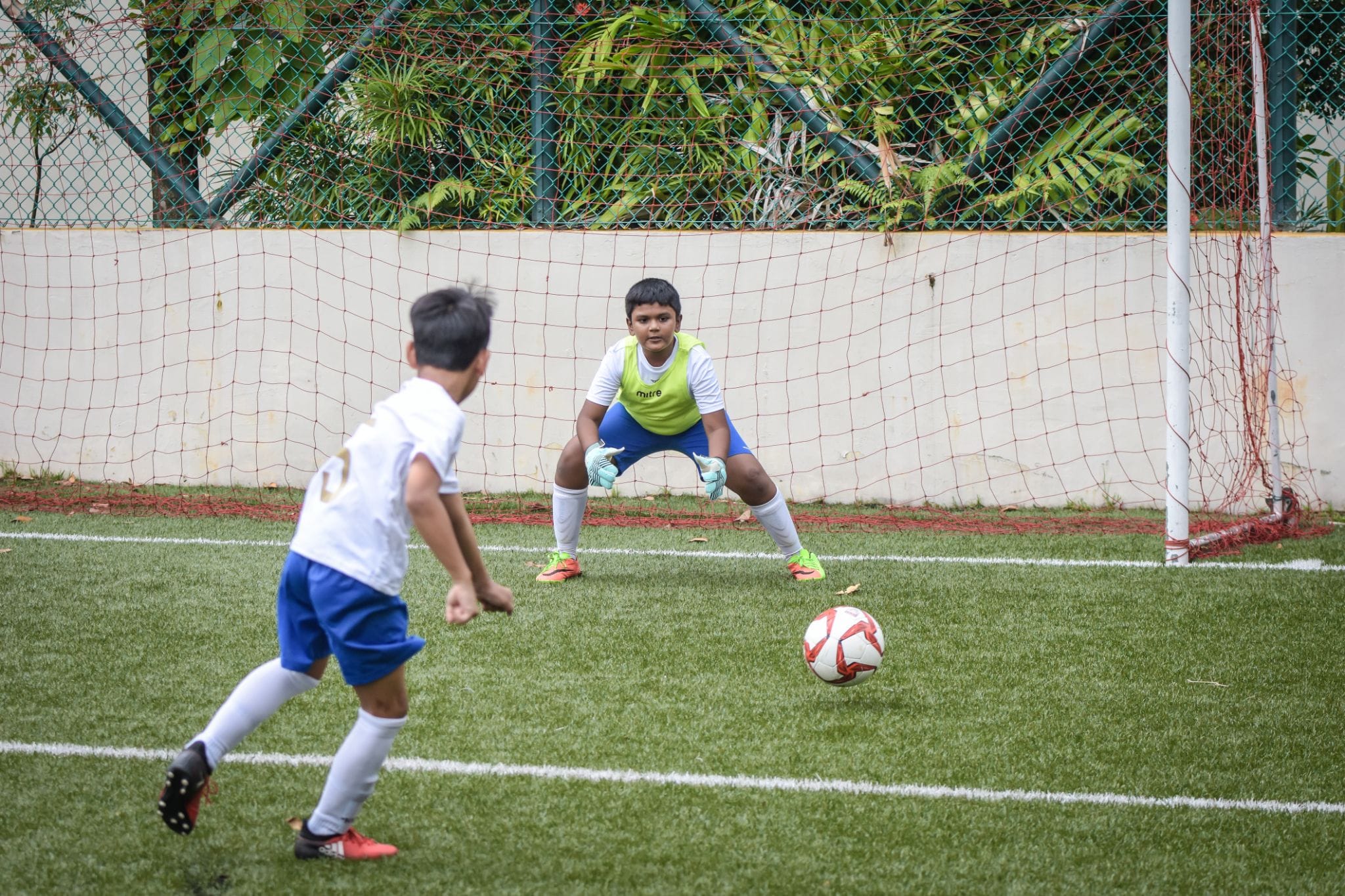 Two young boys playing football, one preparing to shoot while the goalkeeper crouches ready in front of a red net.