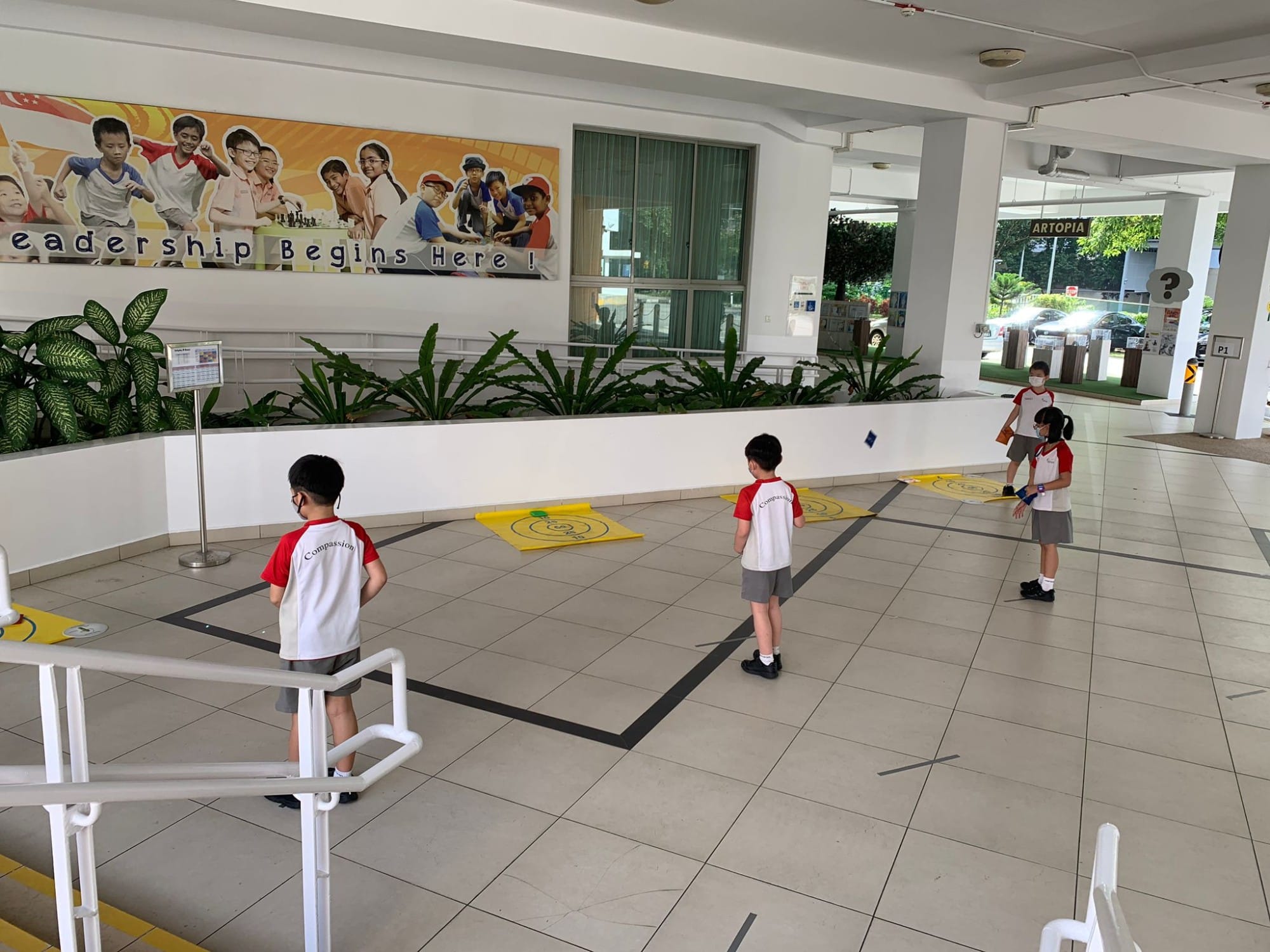 Four masked school children in red and white uniforms stand spaced apart in a covered school corridor with a Leadership Begins Here banner.