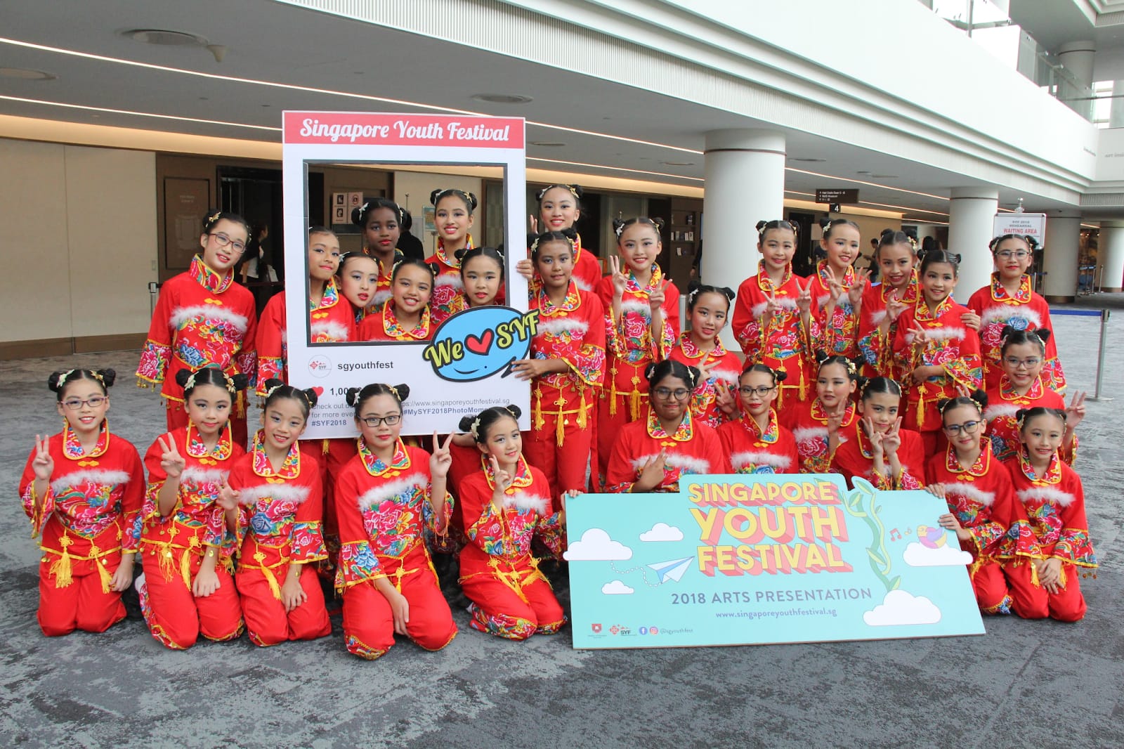 Girls in red Chinese costumes pose with Singapore Youth Festival 2018 Arts Presentation signs indoors.
