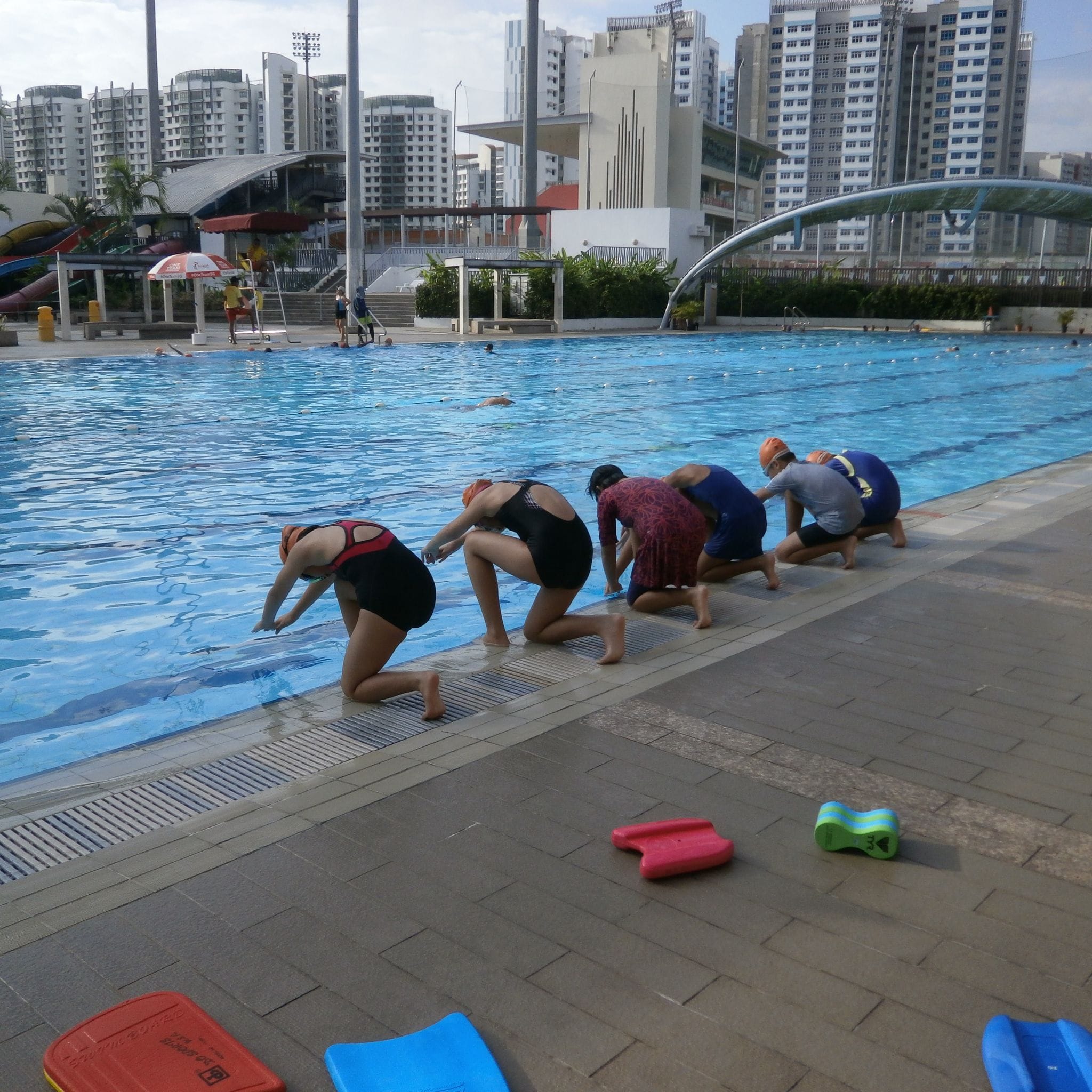 Four swimmers in caps kneel at the edge of an outdoor pool, with kickboards nearby and city buildings behind.
