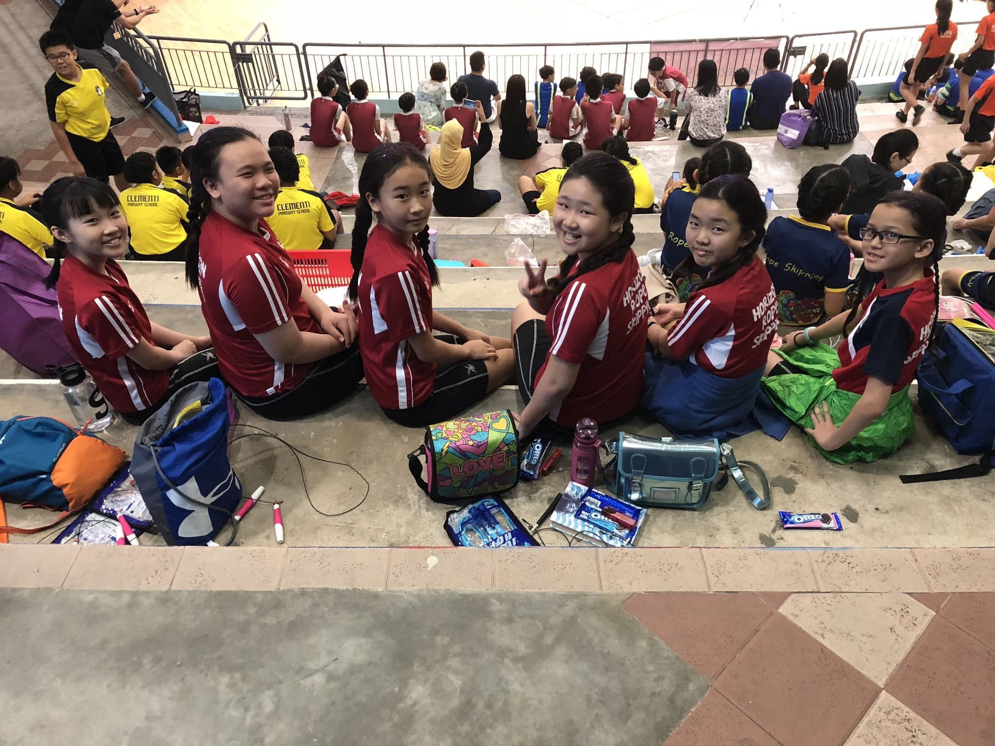 Six girls in red sports uniforms sit on bleachers smiling, with bags and Oreo snacks in front of them.