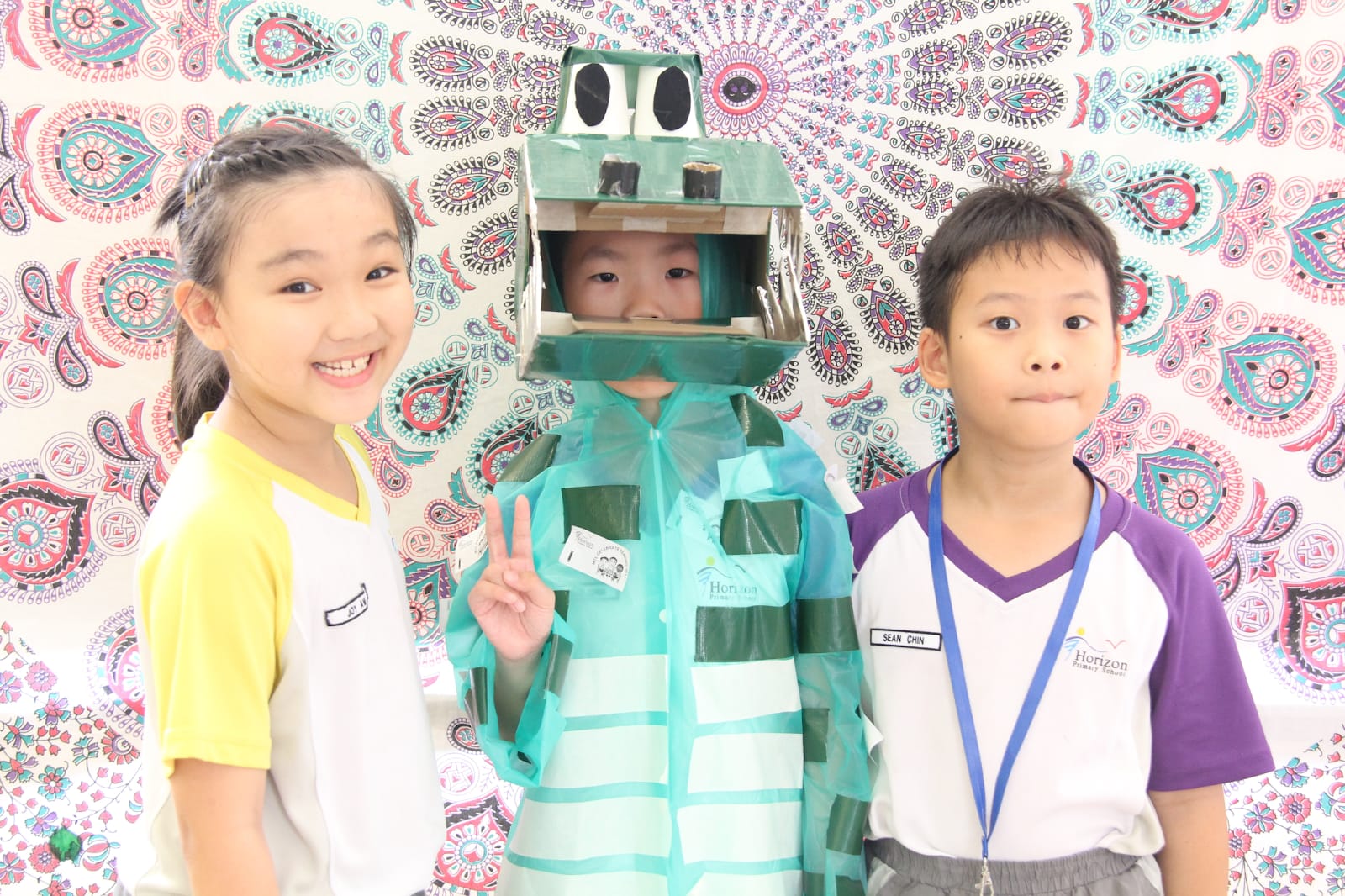 Three children pose together, one wearing a handmade cardboard robot costume, in front of a colorful paisley backdrop.