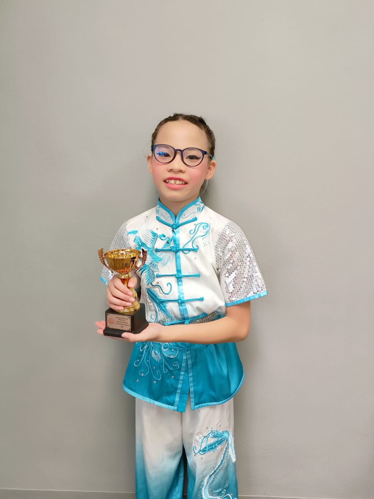 A young girl in a teal and white wushu costume and glasses smiles while holding a gold trophy.